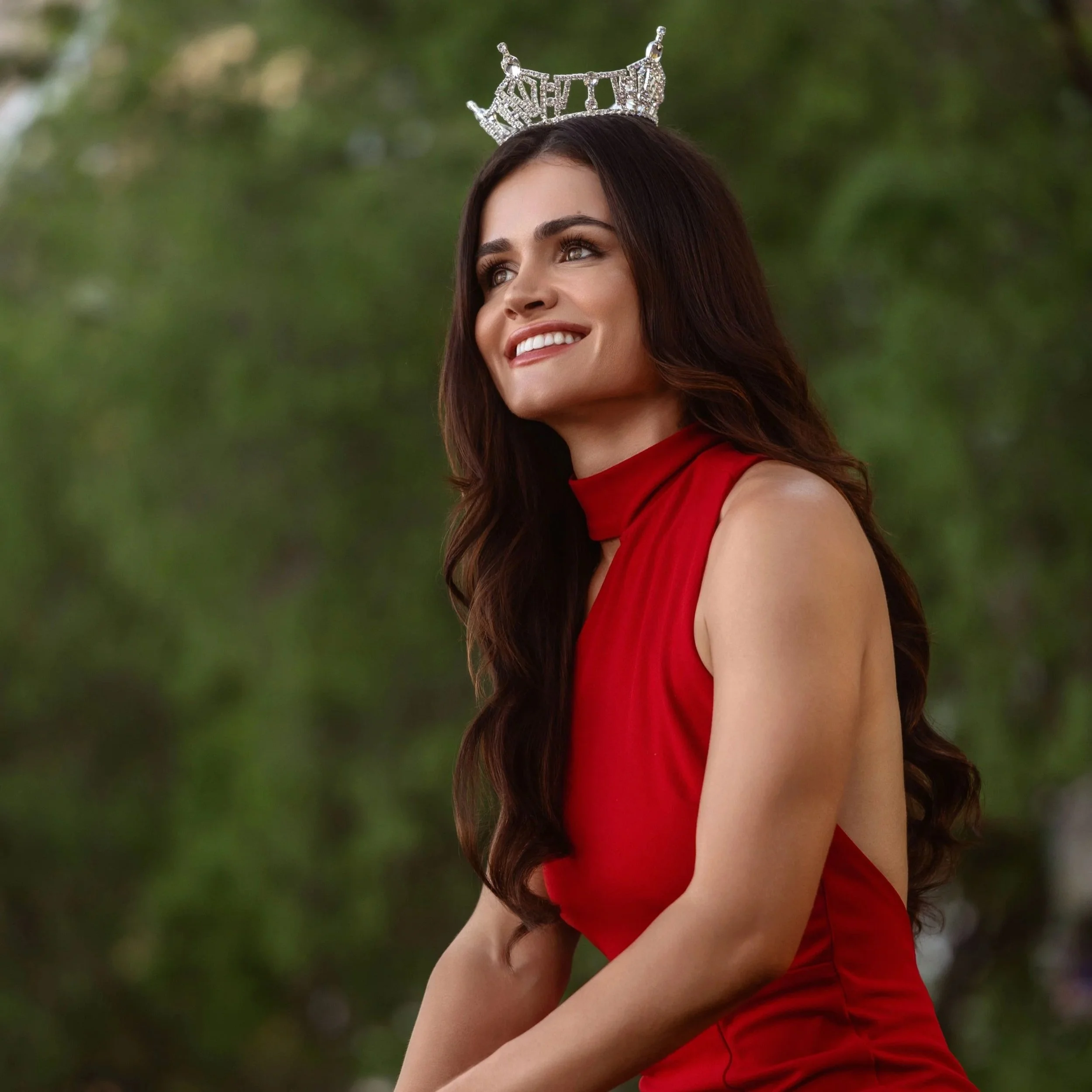 Woman with long dark hair smiling, wearing a silver tiara and a red dress, outdoor background with green trees.