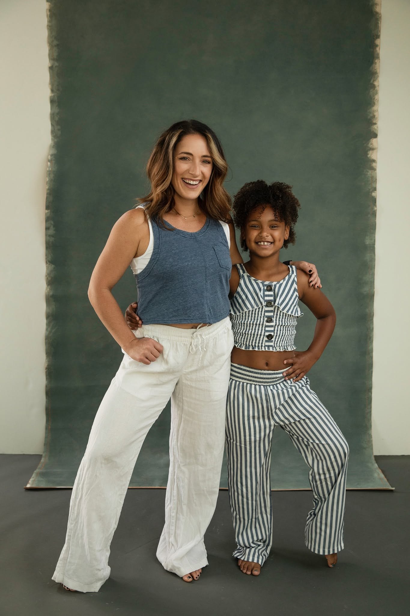 Two women are standing close together, smiling at the camera, with a dark green background behind them. One woman has light brown, shoulder-length hair and is wearing a sleeveless blue top and white pants. The other woman has short, curly hair and is wearing a striped sleeveless top and striped pants.