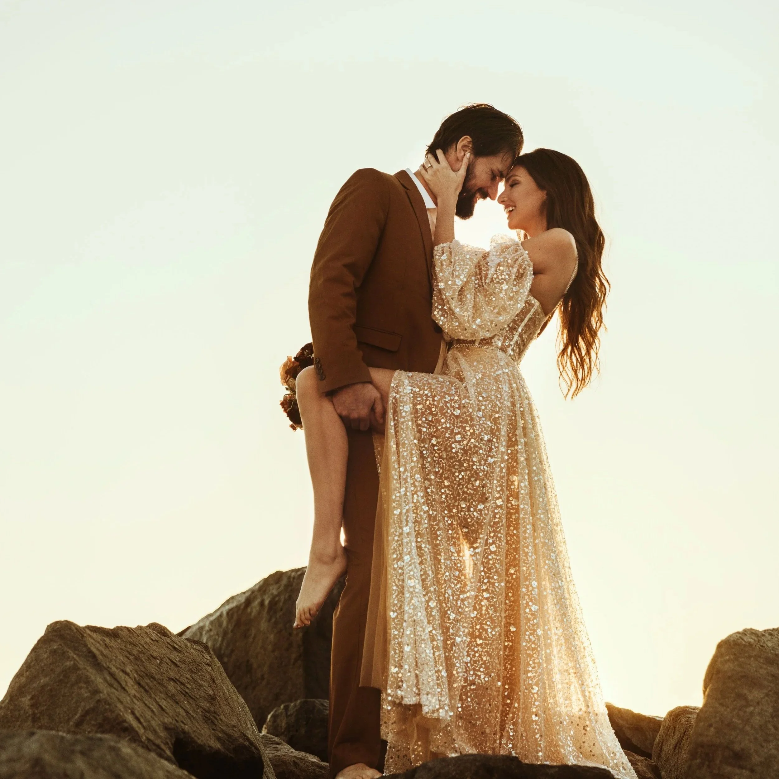 A couple embracing on rocky terrain, the man in a brown suit and the woman in a short, sparkly dress, with a clear sky in the background.