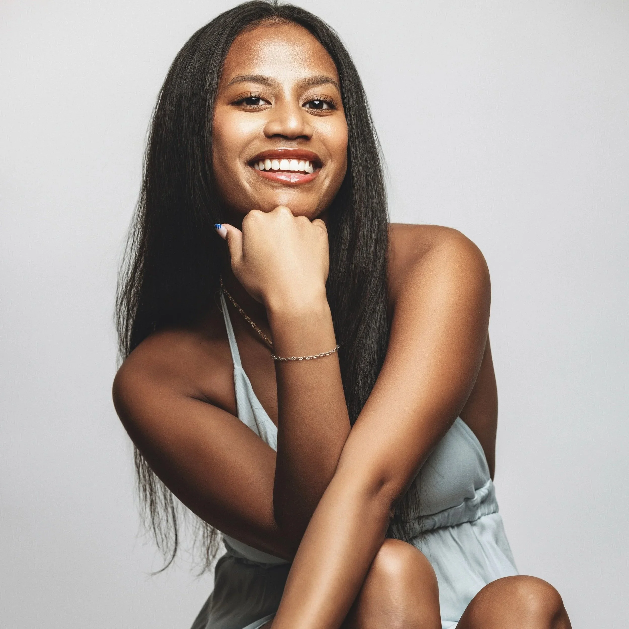 A smiling woman with long black hair, wearing a light-colored top and sitting against a plain light background.