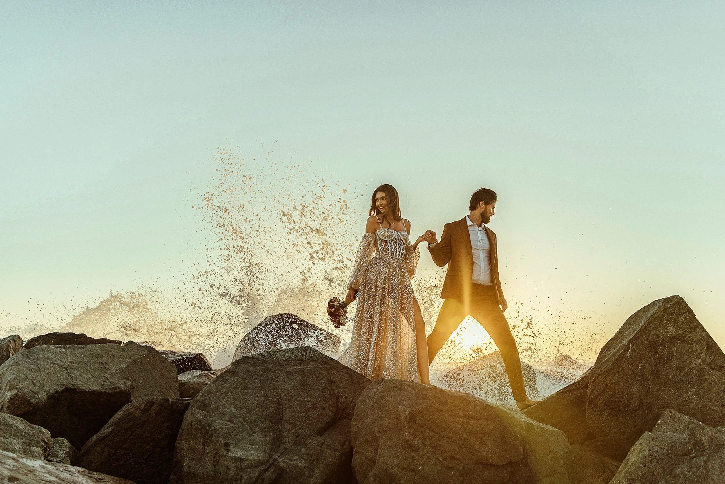 A couple holding hands walking on rocks by the water at sunset, with splashing waves and a clear sky.