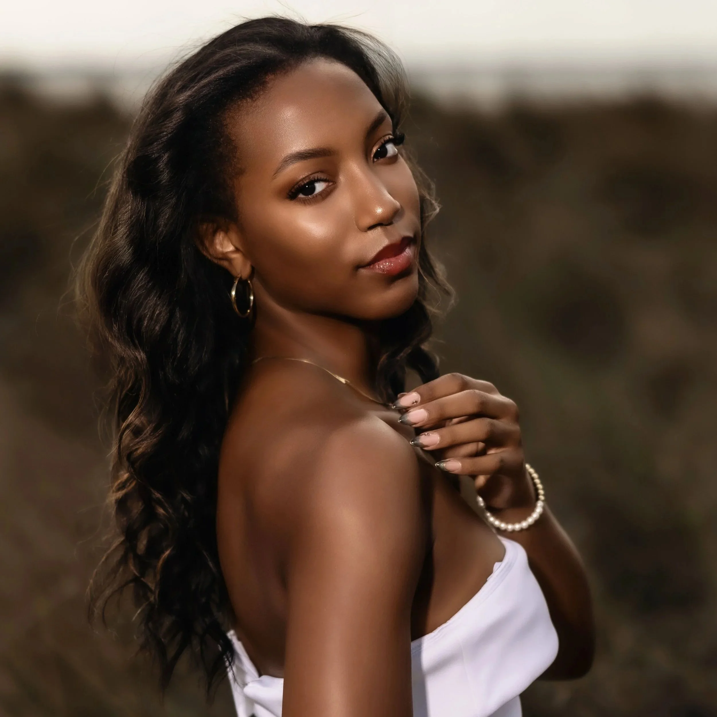 Close-up of a woman with dark, curly hair, wearing gold hoop earrings and a pearl necklace, looking over her shoulder with a confident expression, in an outdoor setting.