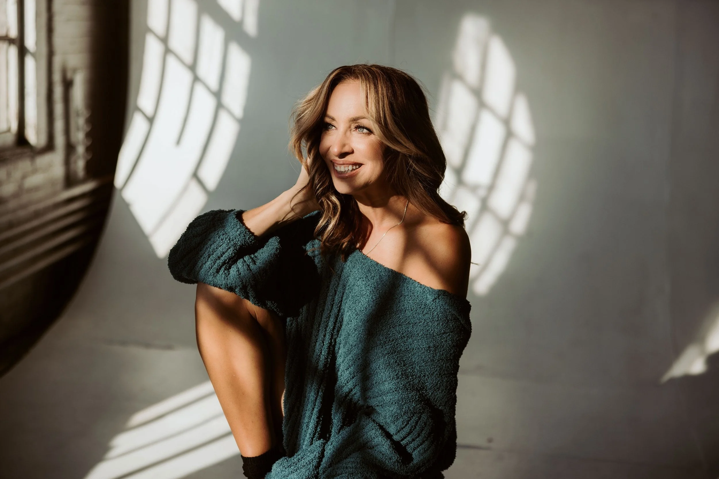 A woman with shoulder-length brown hair, smiling and looking to the side, standing in a room with sunlight casting shadows through window blinds on the wall behind her.