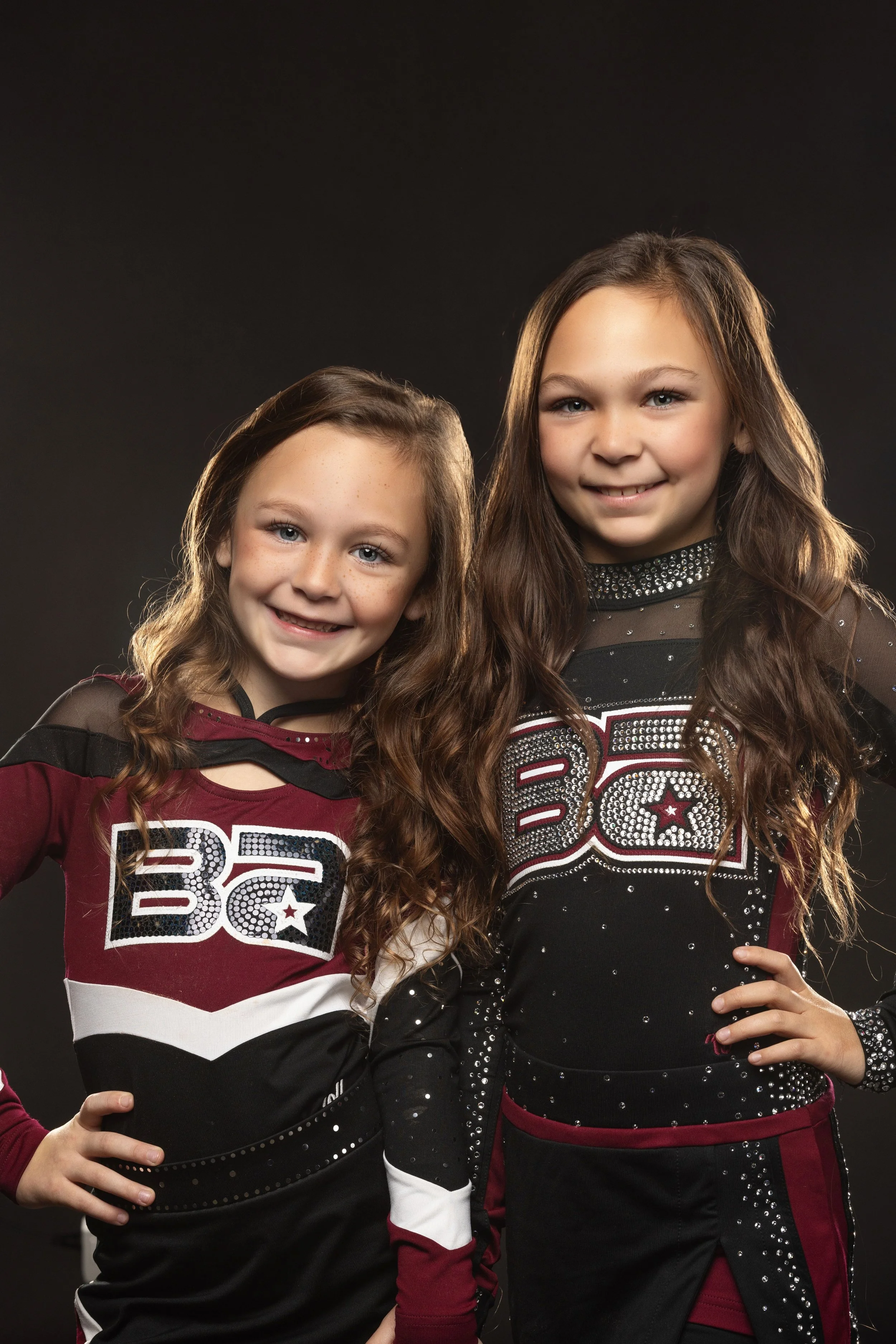 Two young girls dressed in cheerleading uniforms stand against a black background, smiling and posing for the camera.