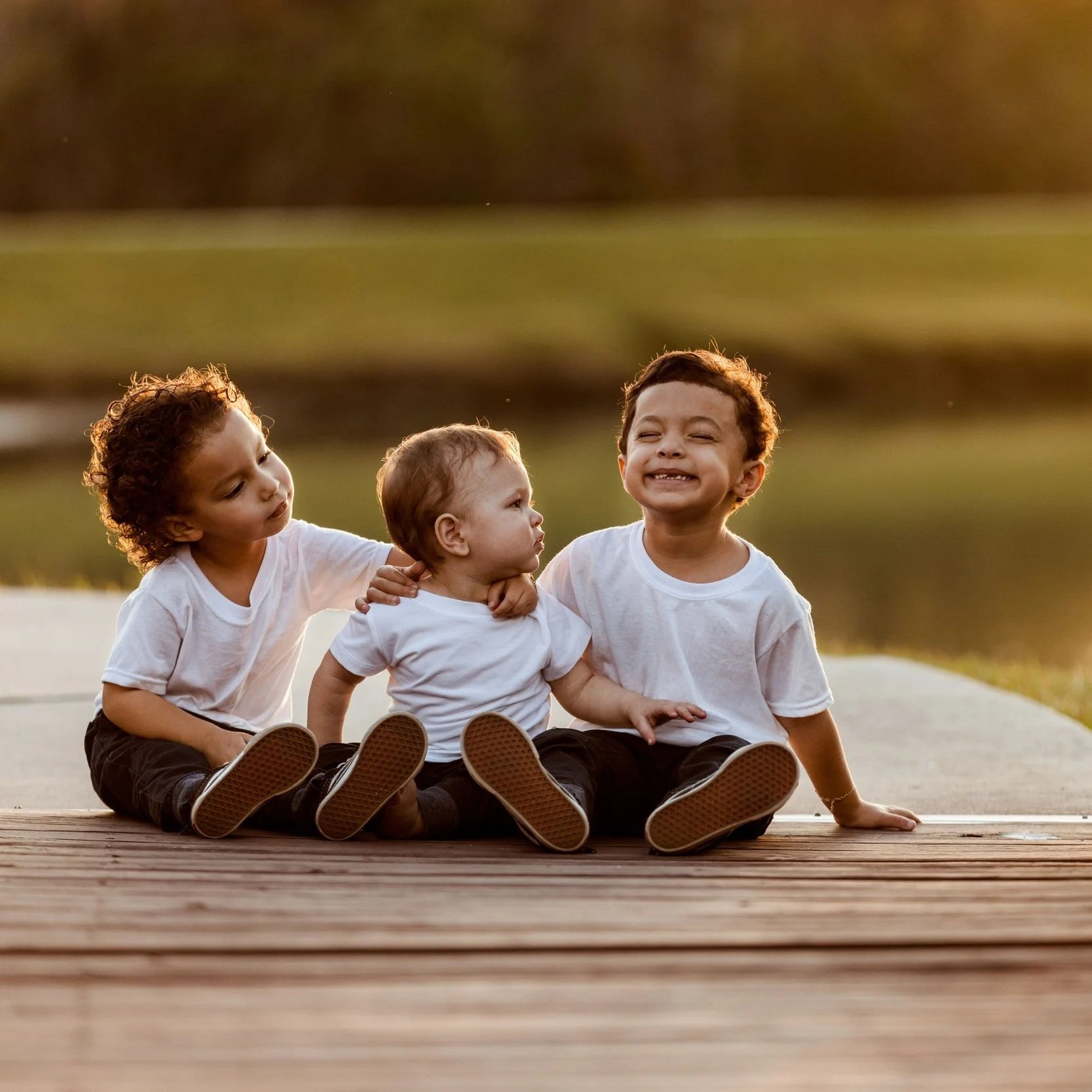 Three children sitting on a wooden deck outdoors with a blurred green field in the background. The children are wearing white shirts and black pants, displaying happy and playful expressions.