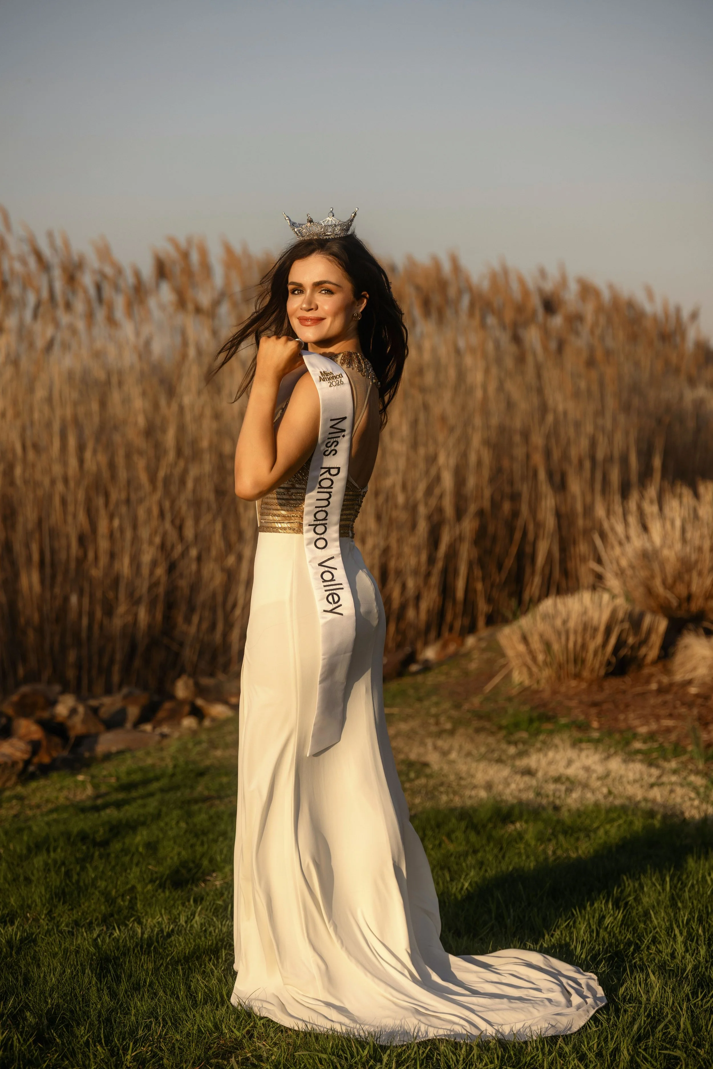 A woman wearing a crown and a sash that says 'Miss Rodeo Valley' stands in a field of tall, dried grass, smiling and looking at the camera during sunset.