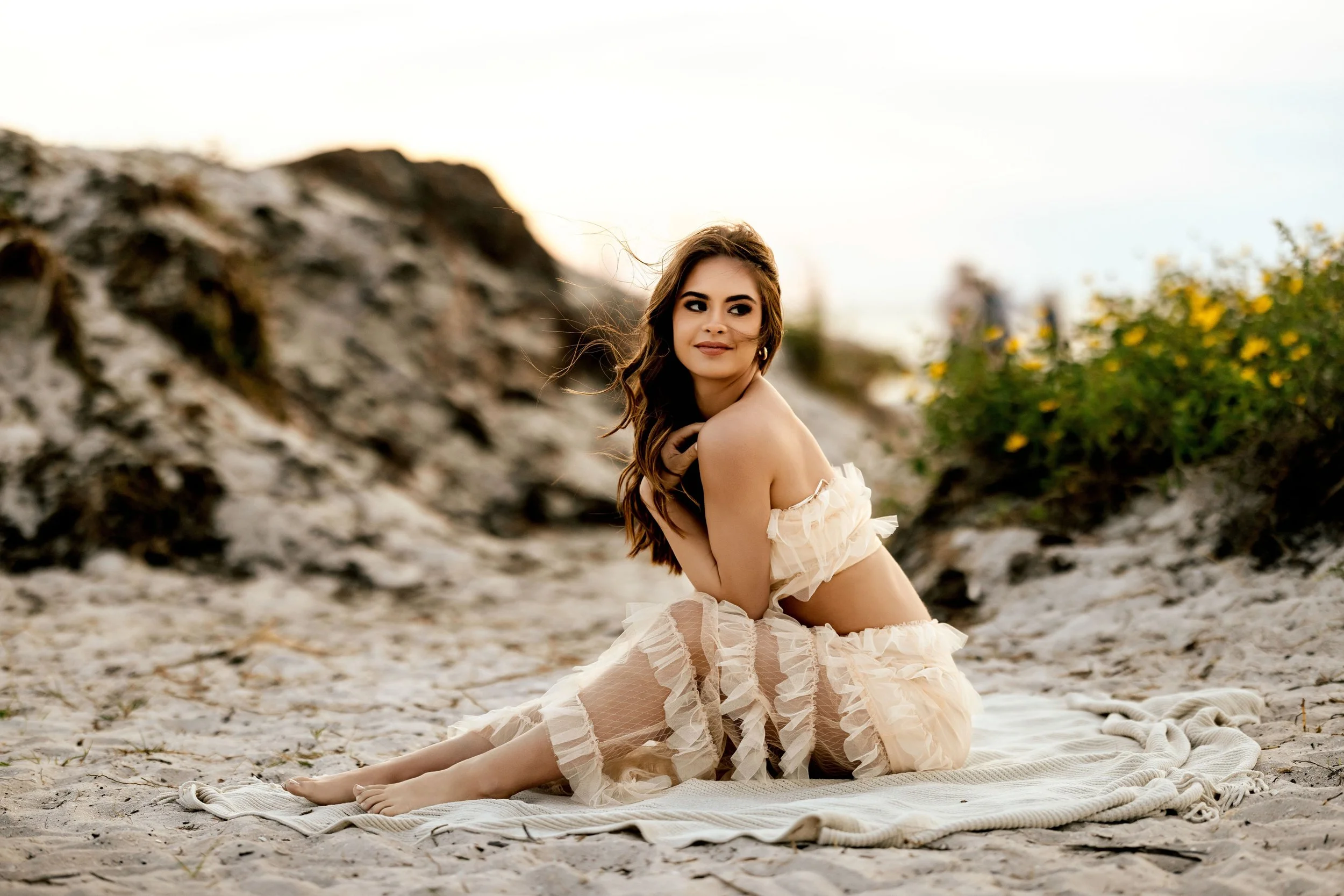 A woman with long brown hair sitting on a sandy beach near rocks and greenery, wearing a ruffled beige outfit, with wind blowing her hair and a relaxed smile.