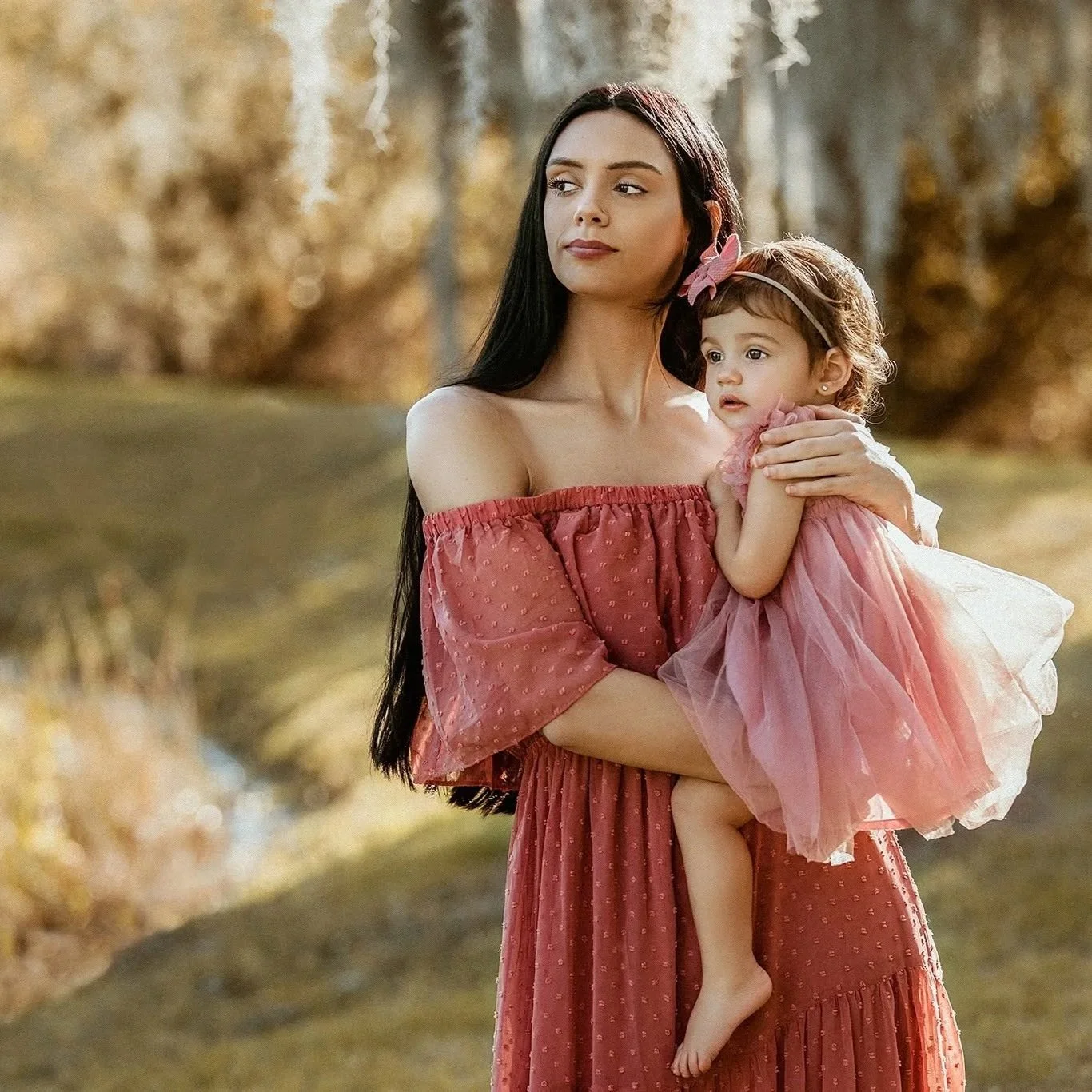 A woman with long dark hair holding a small girl in a pink dress outdoors in a natural setting with trees and a body of water in the background.