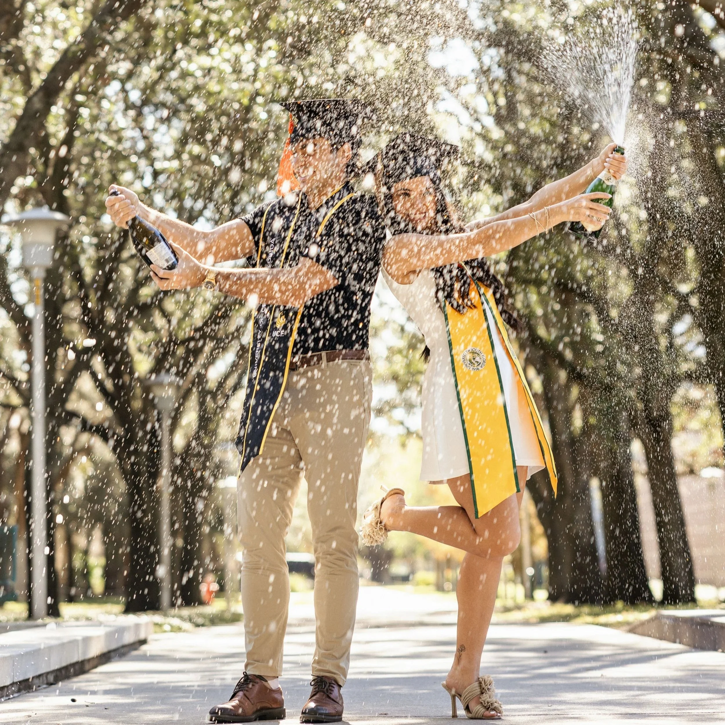 Two people celebrating graduation outdoors, spraying champagne into the air, with trees in the background.