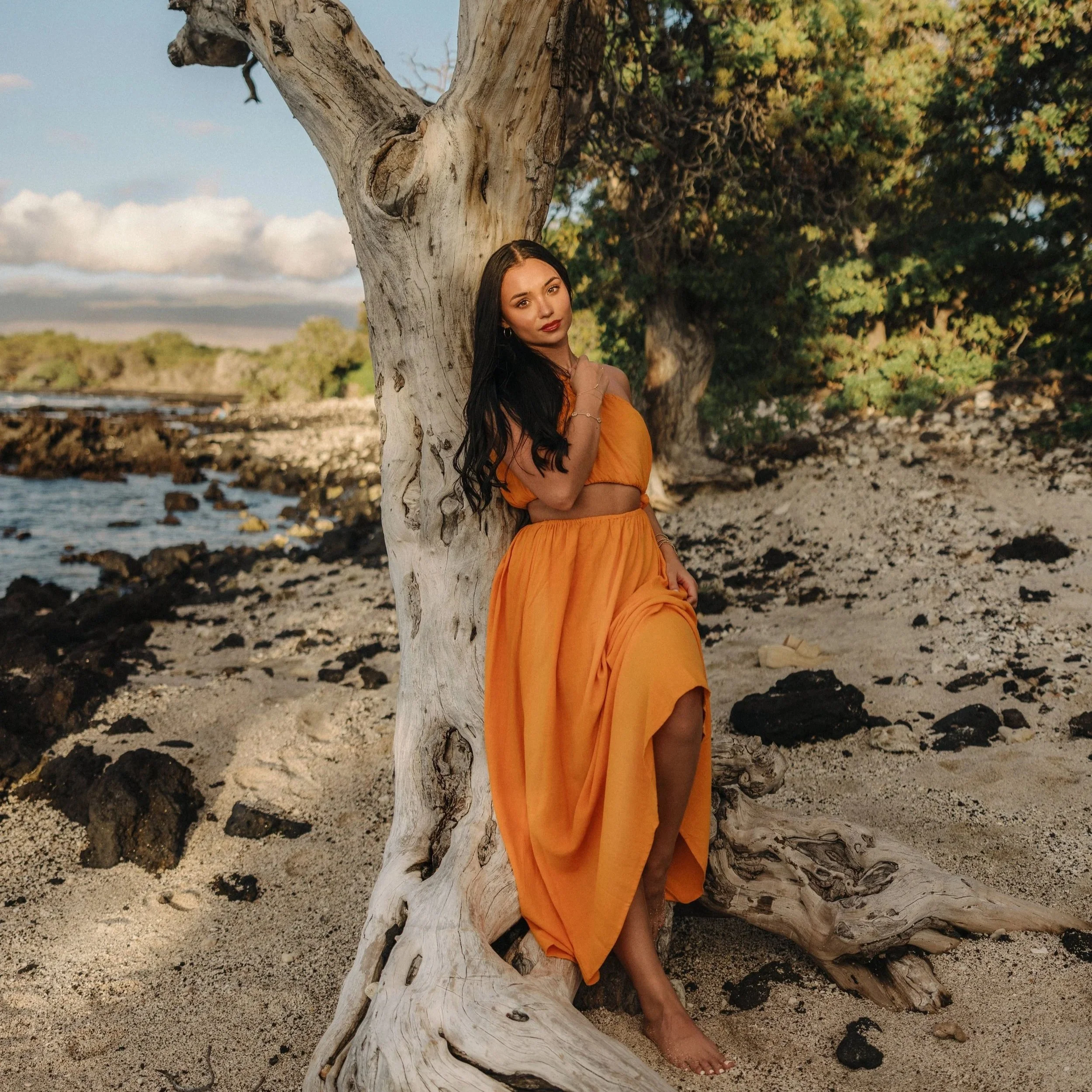 Woman with long dark hair wearing an orange off-shoulder top and matching skirt, standing behind a large twisted driftwood log on a sandy beach with a rocky shoreline and green trees in the background, under a partly cloudy sky.