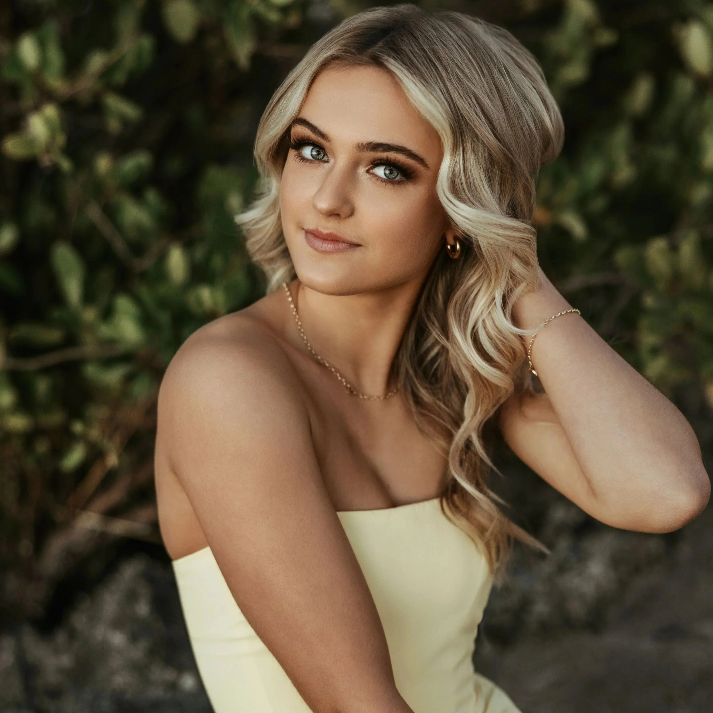 A woman with blonde hair styled in loose waves, dressed in a strapless yellow top, standing outdoors against a blurred green foliage background.