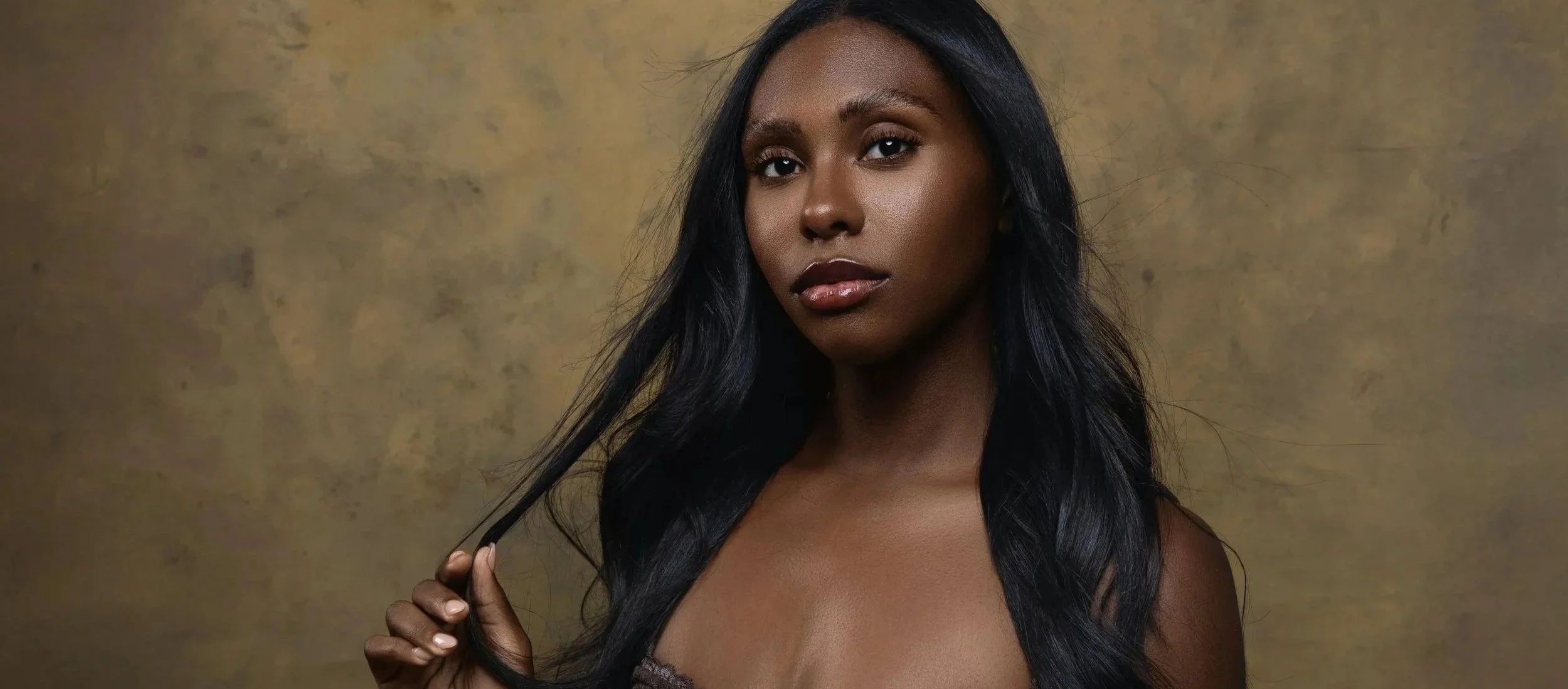 A woman with long black hair, dark skin, and full lips, looking at the camera, holding her hair with her right hand, against a textured beige background.