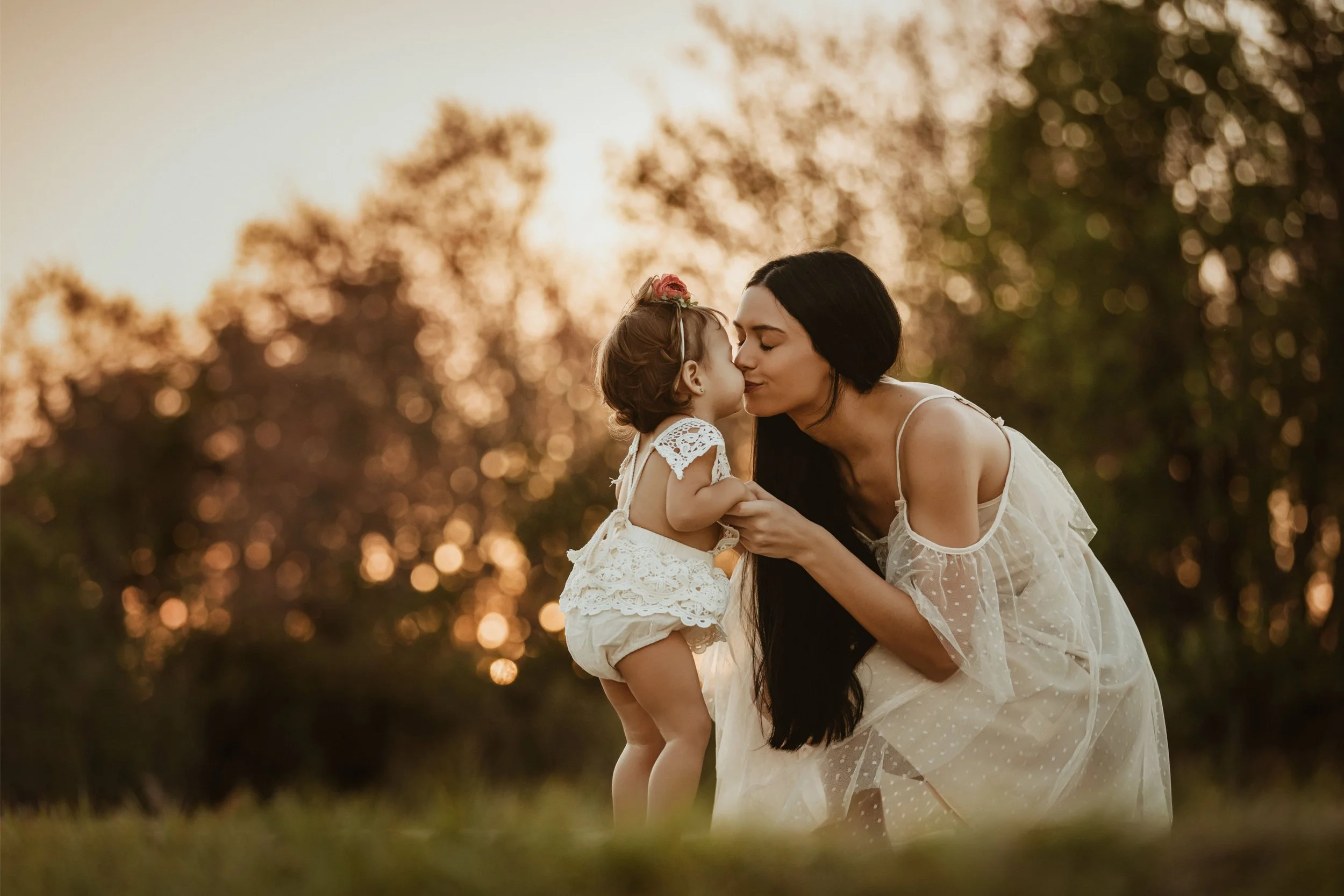 A woman and a young girl share a kiss outdoors during sunset, with blurred trees and warm sunlight in the background.