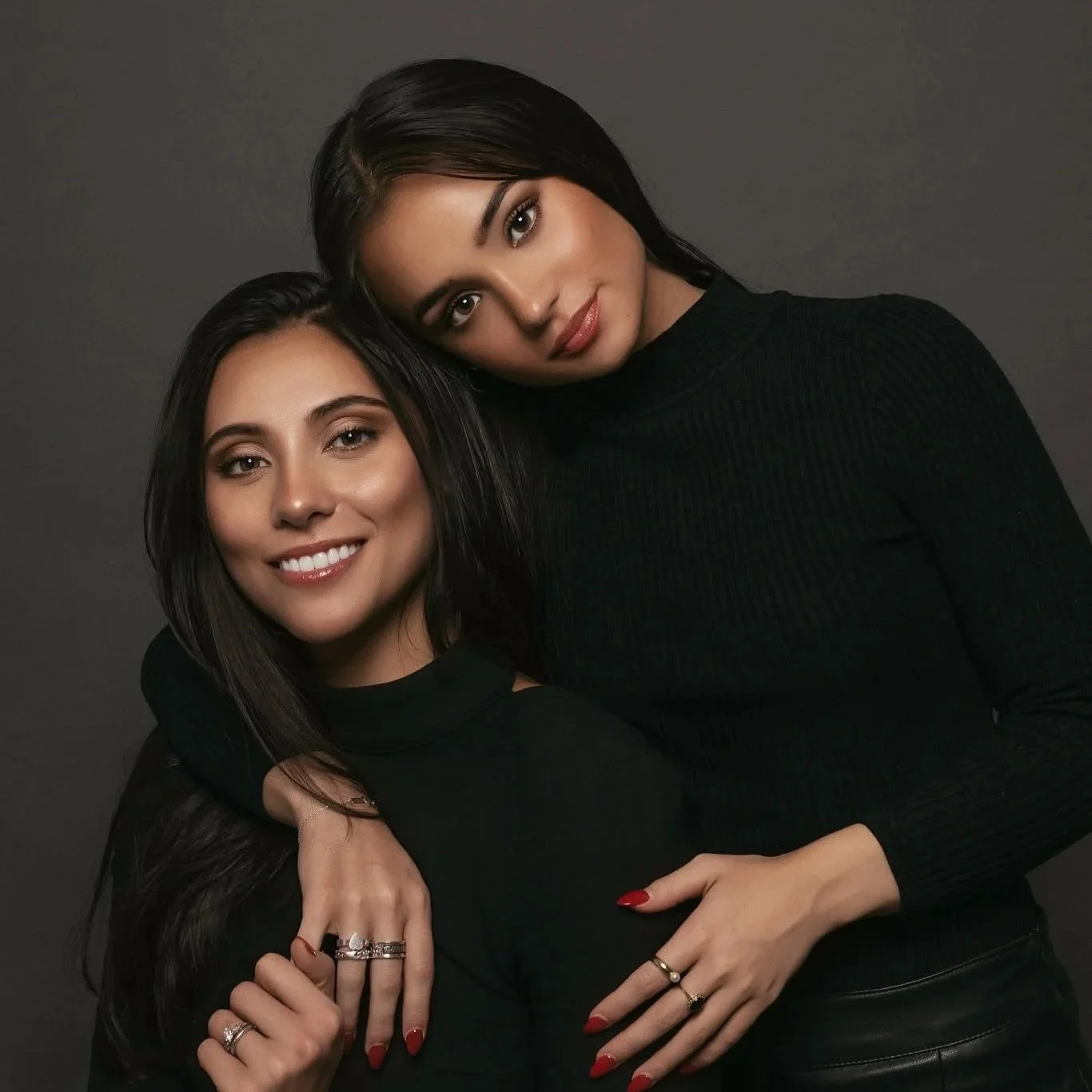 Two women hugging and smiling, both with dark hair and black clothing, against a dark gray background.