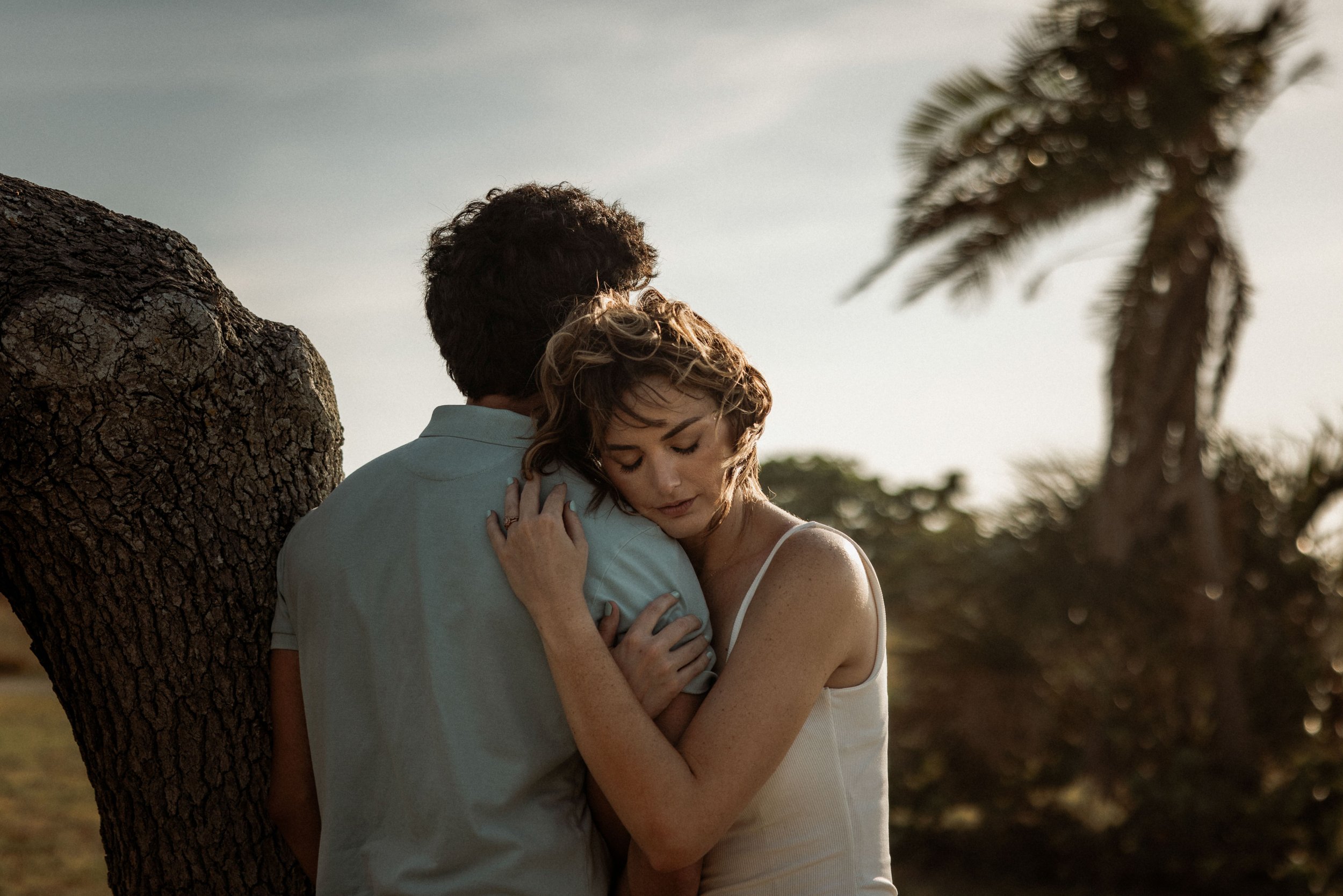 A couple embracing outdoors near a tree during sunset, with a palm tree in the background.