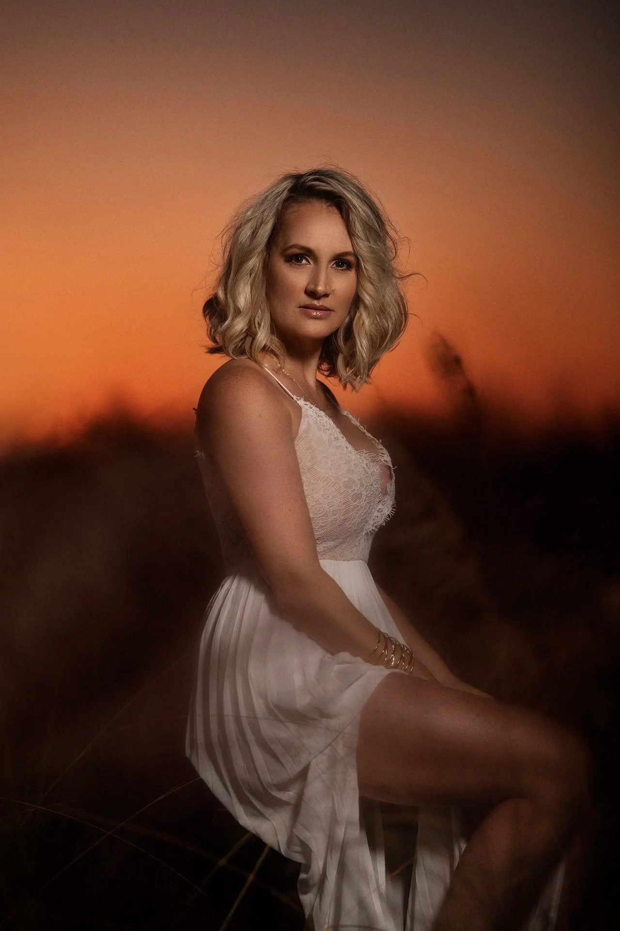 A woman with blonde, shoulder-length curly hair sitting outdoors during sunset, wearing a white lace top and a striped white skirt, with a warm, orange sky in the background.
