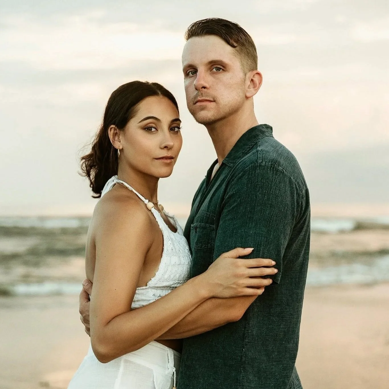 A young couple standing closely on the beach, embracing, with the ocean and sky in the background during sunset.