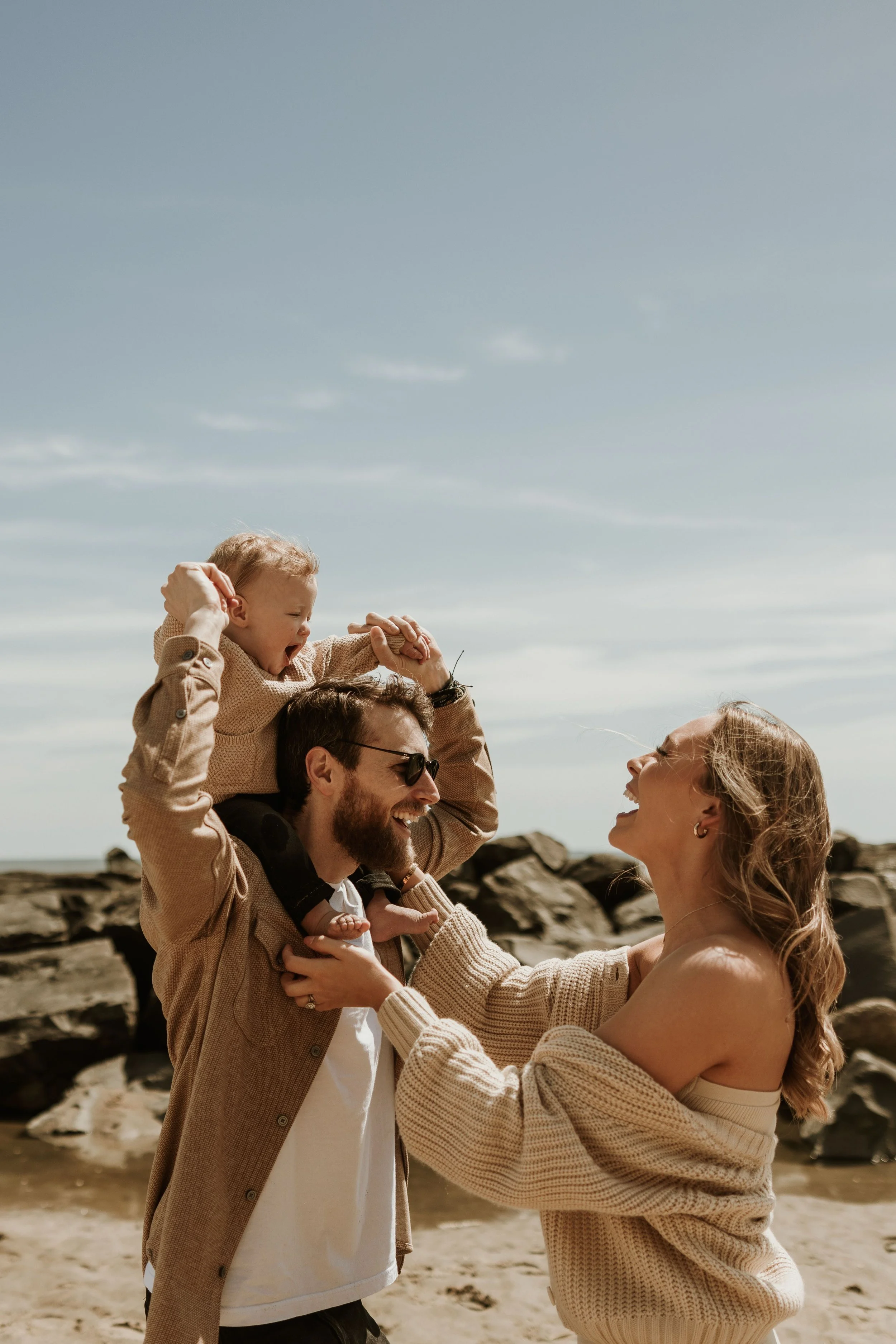 A family at the beach with a man carrying a child on his shoulders, and a woman smiling at the child while holding her hands. Rocks and the sea are visible in the background.