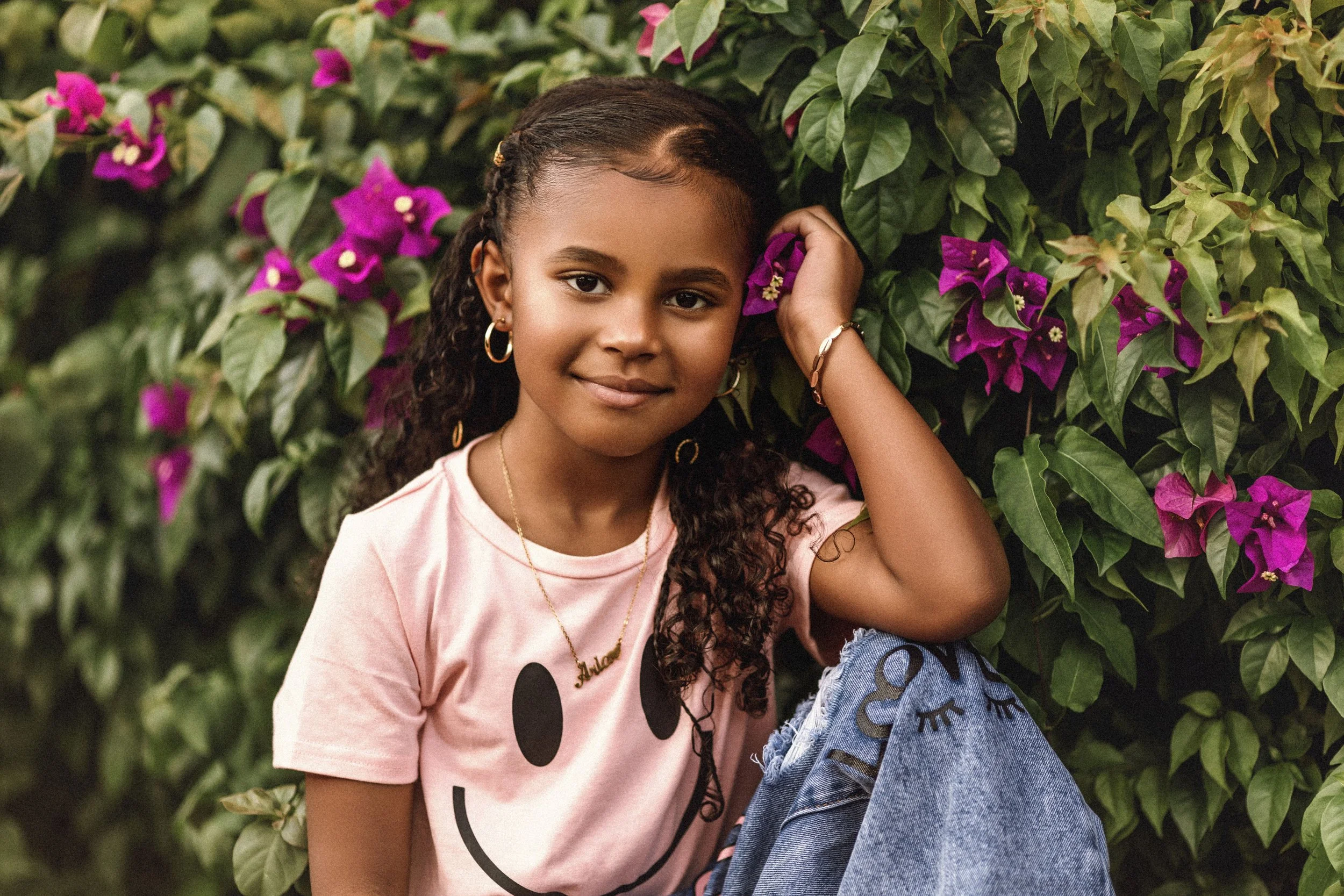 A young girl with dark curly hair, wearing a pink shirt with a smiling face design, gold hoop earrings, and a necklace, is sitting in front of green leafy plants with pink flowers. She is touching her hair and looking at the camera.