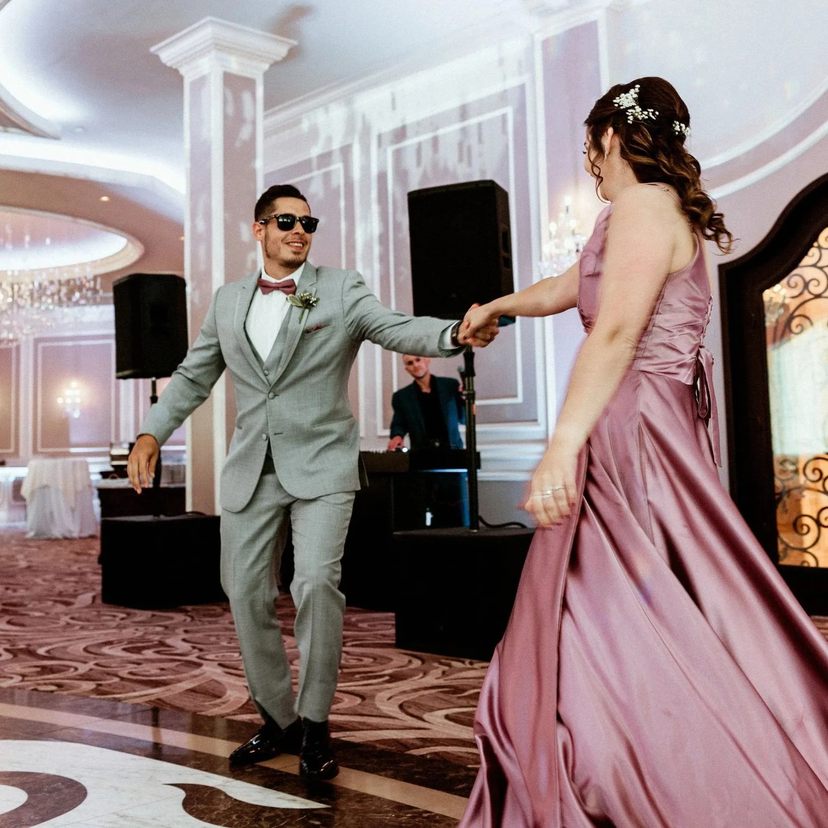 A man in sunglasses and a gray suit with a red bowtie dancing with a woman in a pink satin dress in a lavish ballroom with chandeliers.