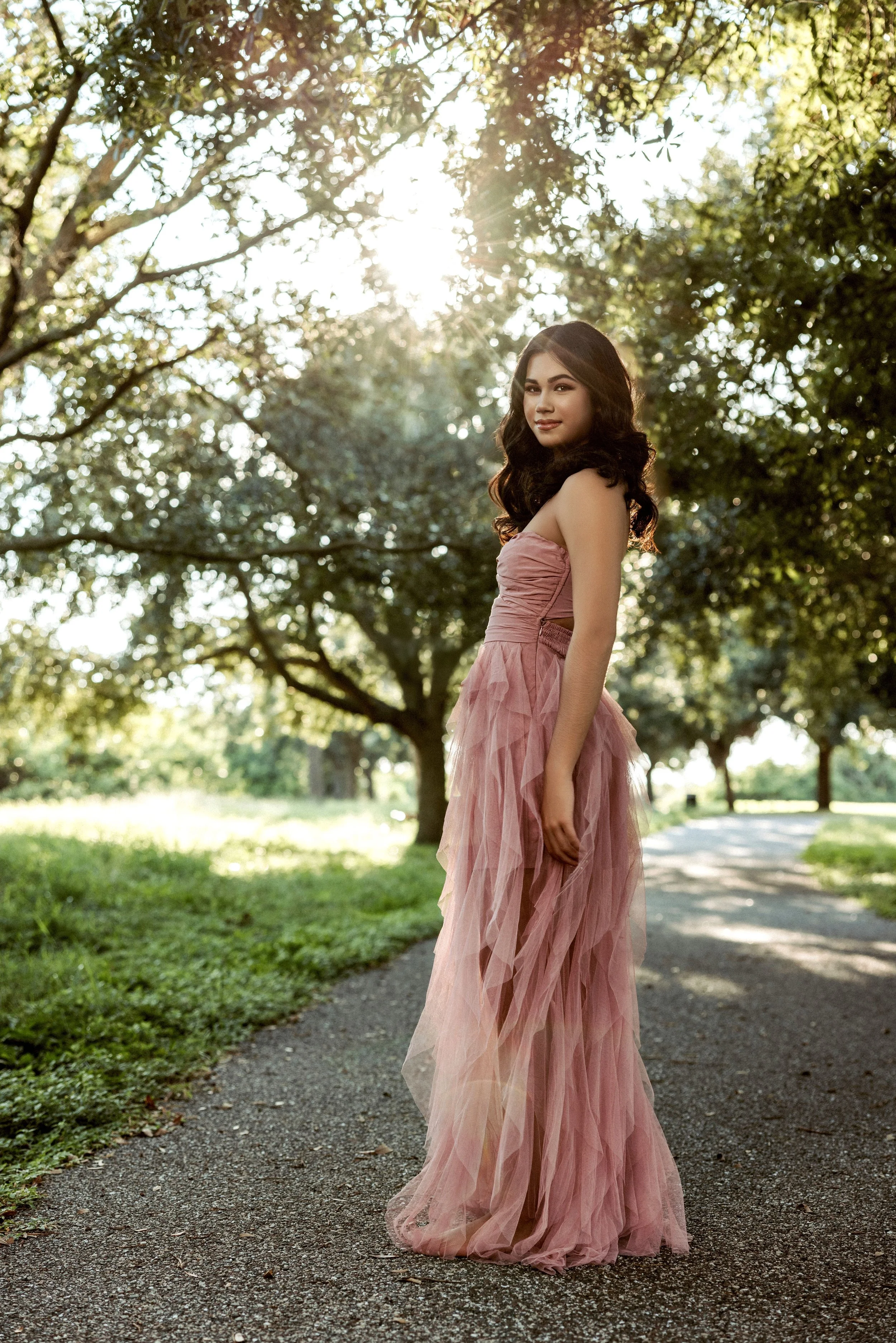 A woman in a pink strapless gown standing on a park pathway with trees and sunlight in the background.