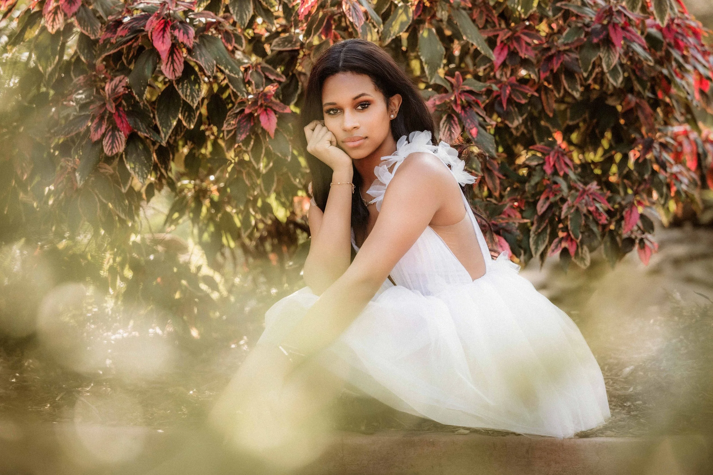 A young woman with dark hair, wearing a white dress with ruffled shoulder details, sitting outdoors in front of a bush with red and green leaves, resting her chin on her hand.