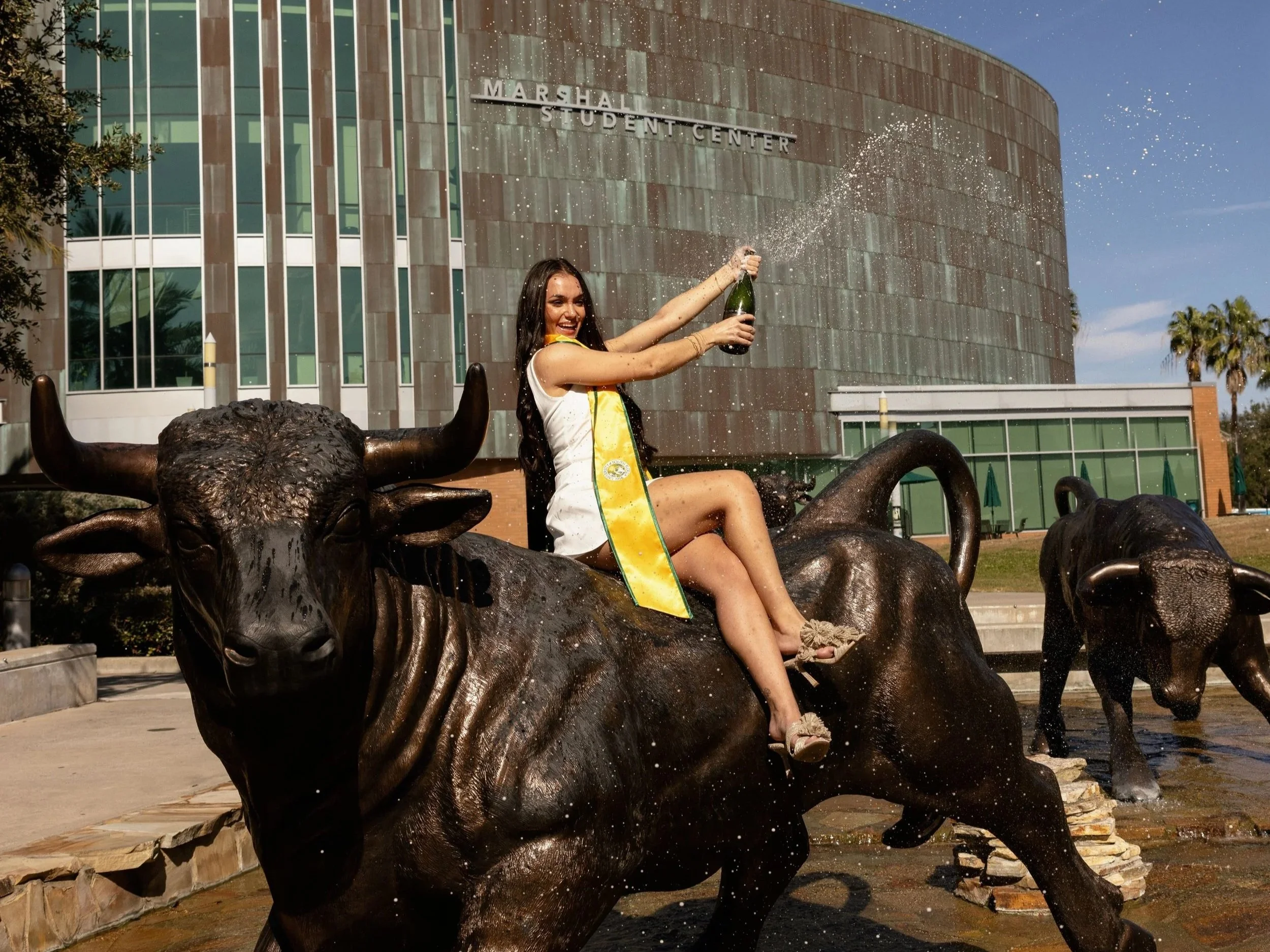 A woman sitting on a bronze sculpture of a bull, holding a bottle of champagne and spraying it, outside the Marshall Student Center building.
