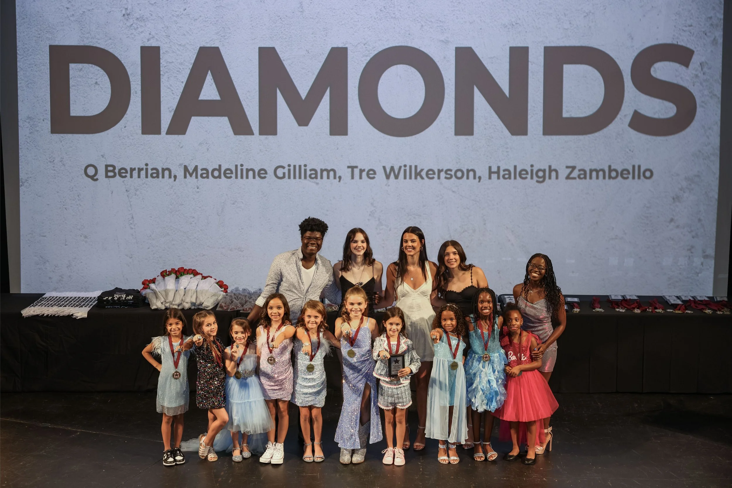 Group of children and adults posing on stage with large screen behind them that says 'DIAMONDS' and a list of names. Some children are wearing medals and fancy dresses, suggesting a celebration or pageant event.