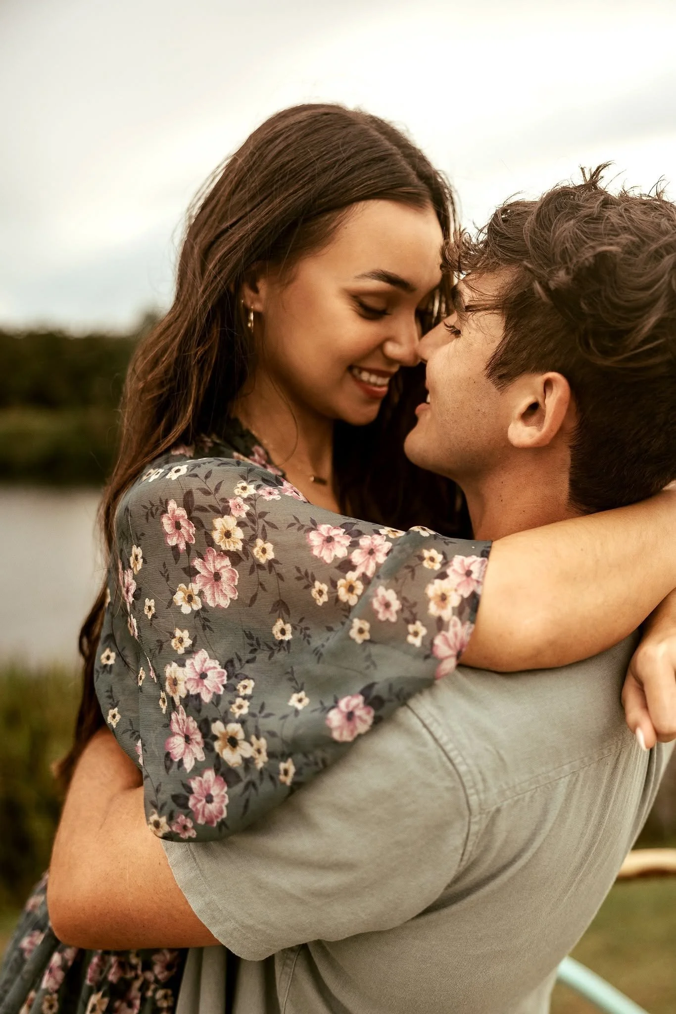 A young woman with long brown hair wearing earrings and a floral dress is smiling with her eyes closed, leaning close to a young man with curly hair, both outdoors near a body of water.