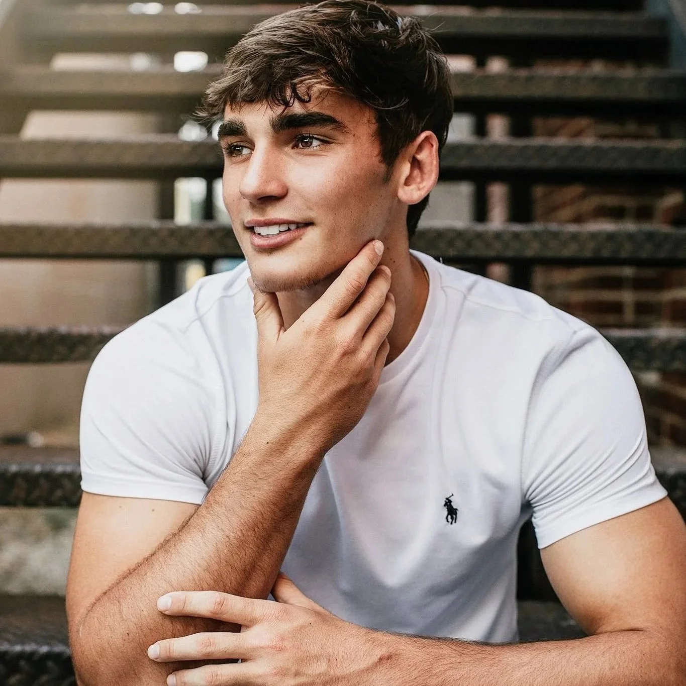 Young man with dark hair and a light complexion sitting outdoors on steps, wearing a white t-shirt with a small logo and touching his neck with one hand while smiling and looking off to the side.