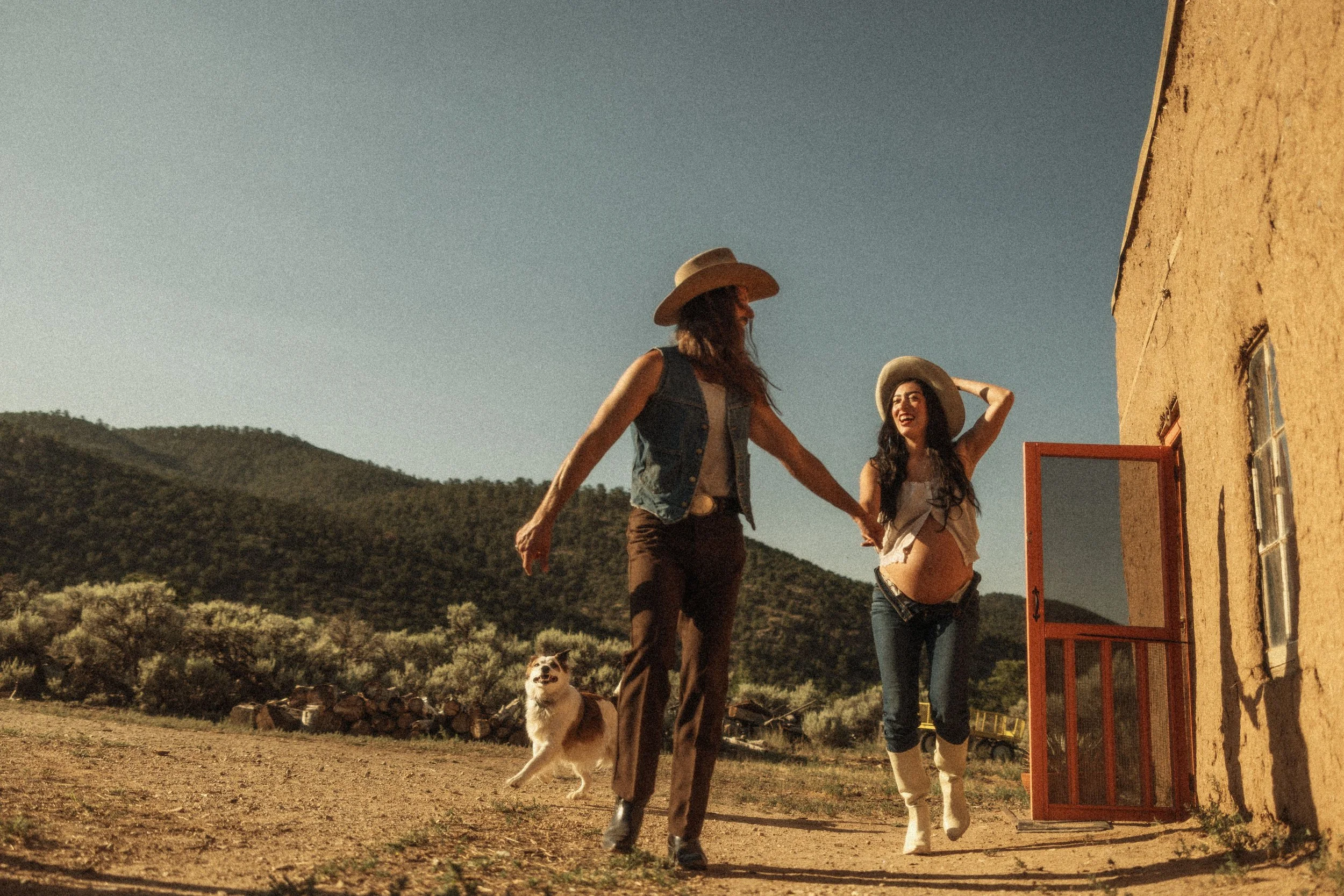 Two women, one with a wide-brimmed hat and denim vest and the other with a lighter hat and cropped top, walk with a dog in a rural outdoor setting near a yellow building with a red door. The background features hills and a clear sky.