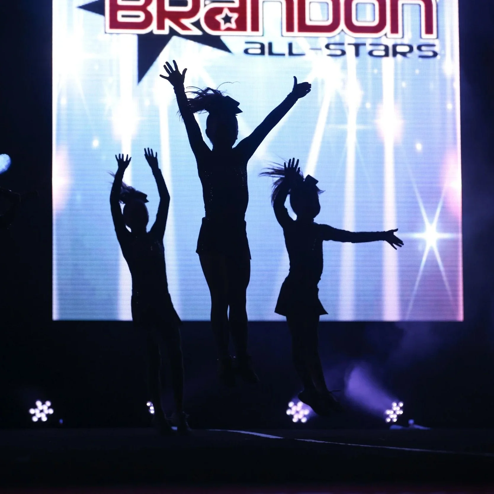 Silhouettes of four dancers performing on stage with a bright LED background displaying the words 'BRONX ALL-STARS' and a star symbol. The stage is dark with spotlights illuminating the performers.