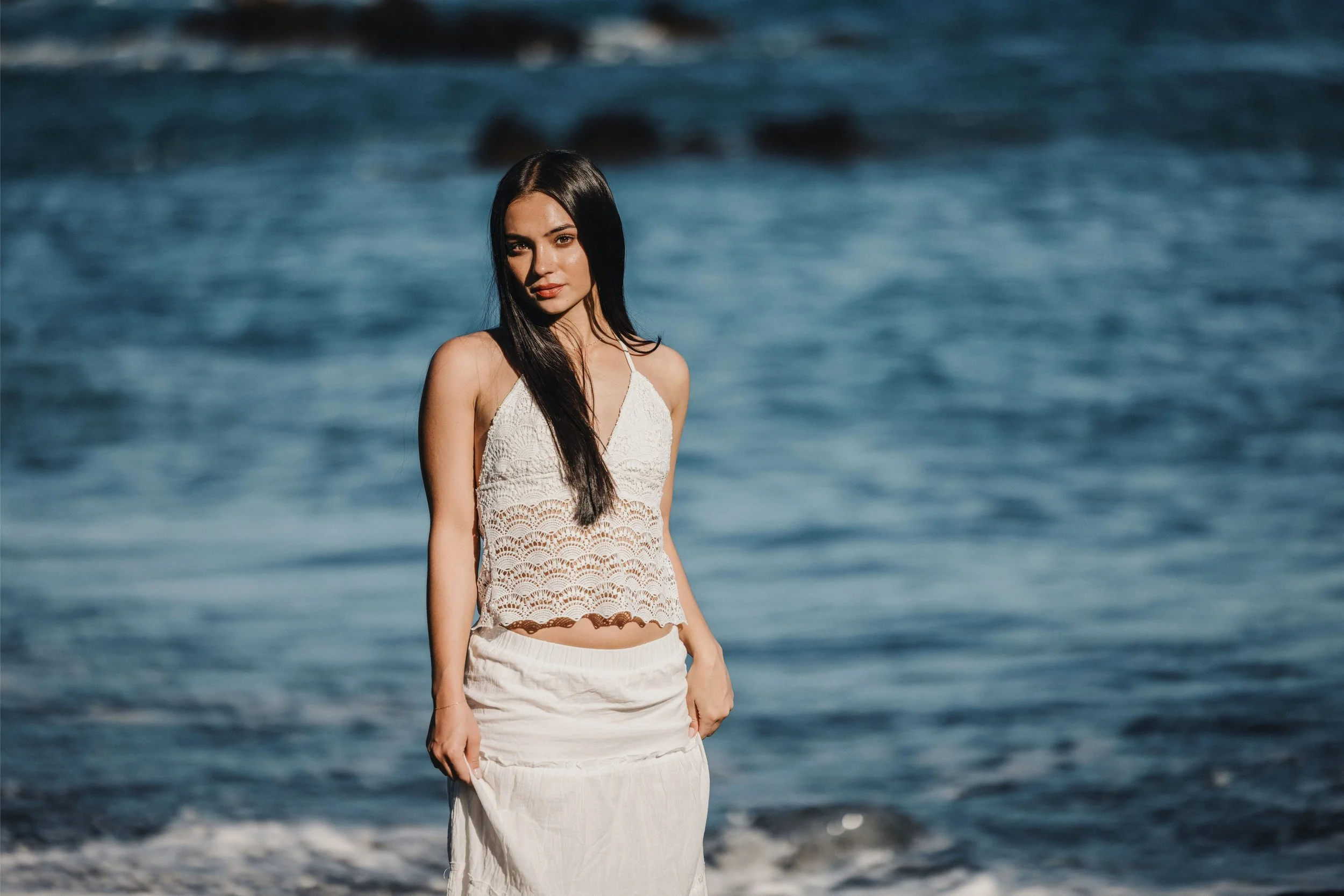 A young woman with long dark hair stands on a beach near the ocean, wearing a white sleeveless lace top and a white skirt, with ocean waves in the background.