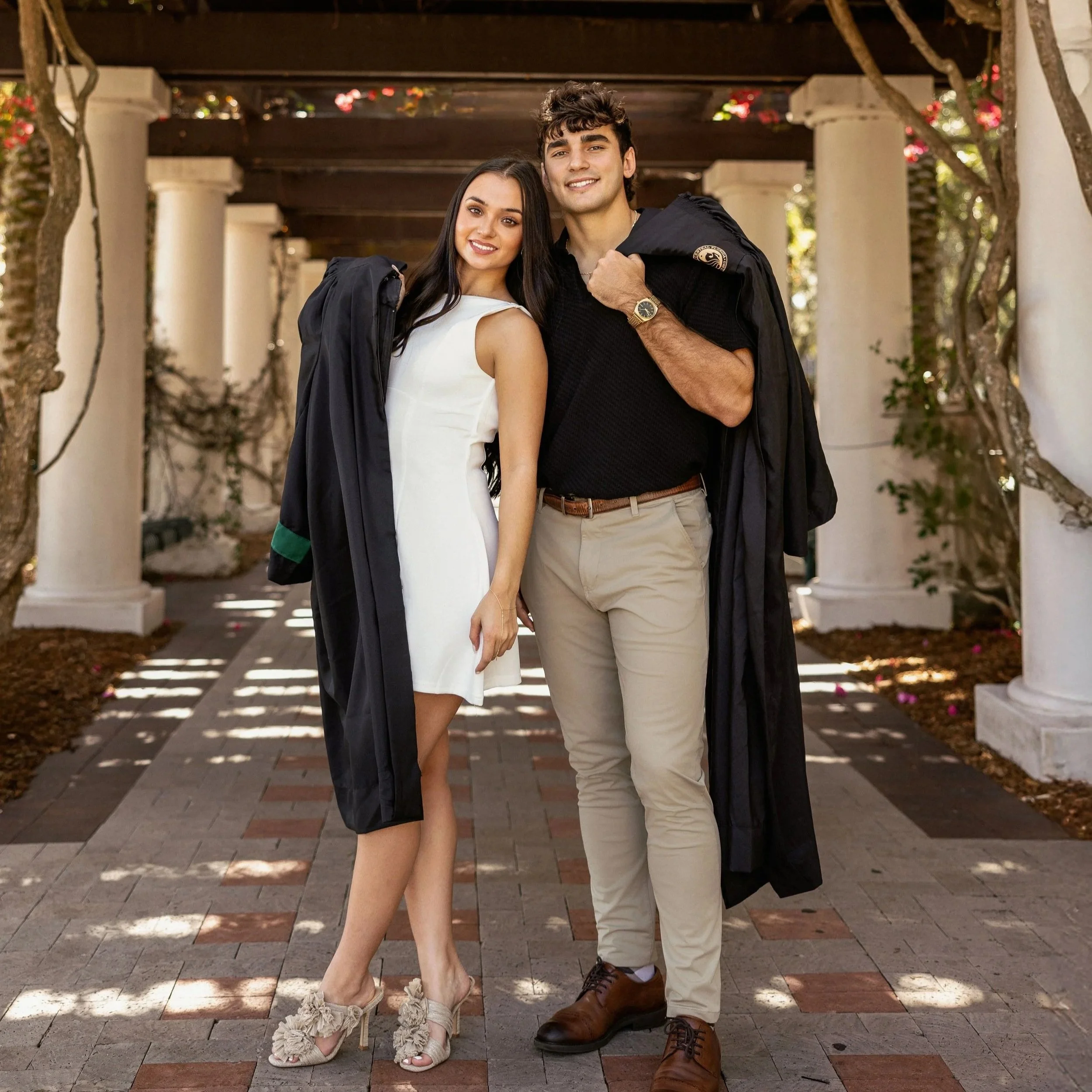 A young woman in a white dress and a young man in a black shirt with beige pants are standing close under a wooden pergola with columns, surrounded by trees and pink flowers, smiling at the camera, holding graduation gowns.