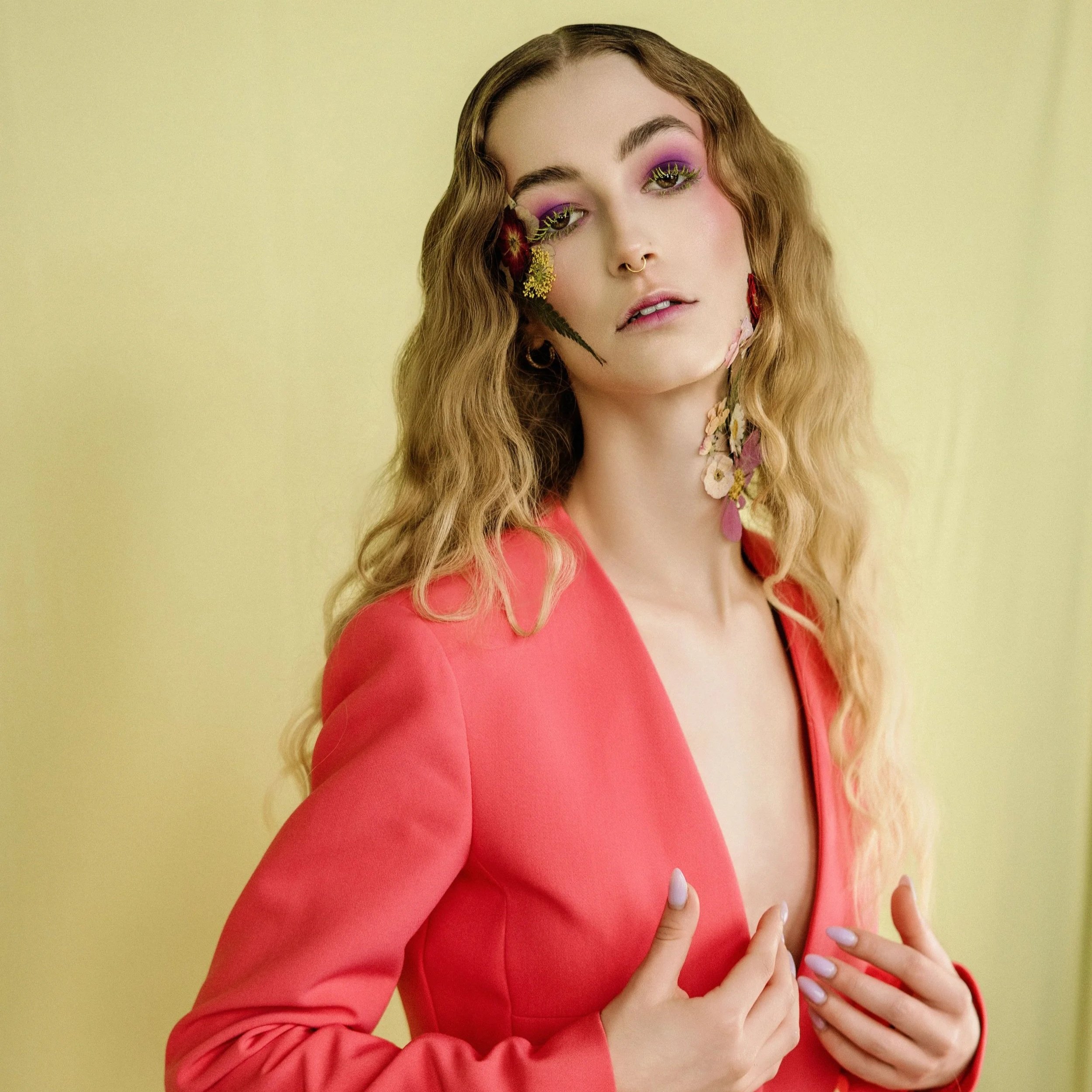 A young woman with wavy light brown hair wearing a pink blazer and colorful makeup, standing against a light yellow background, with her hand on her chest.