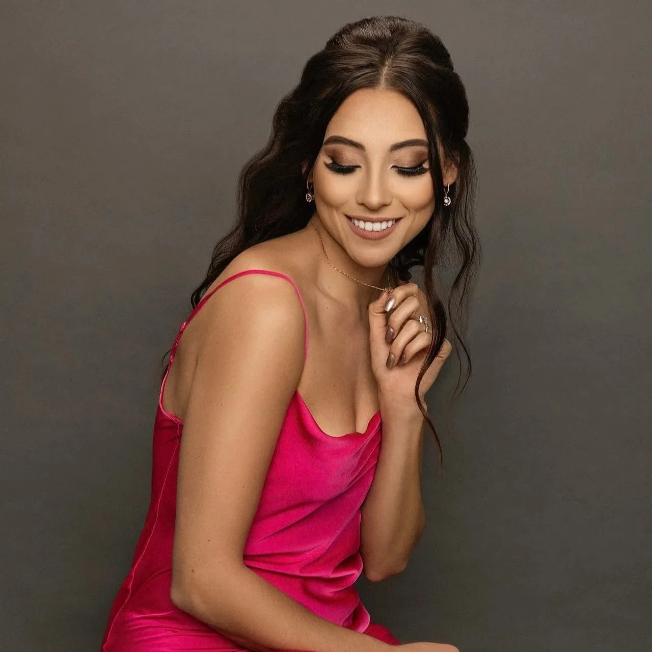 A young woman with long dark hair, wearing a pink satin dress, smiling with her eyes closed, and jewelry, against a plain gray background.