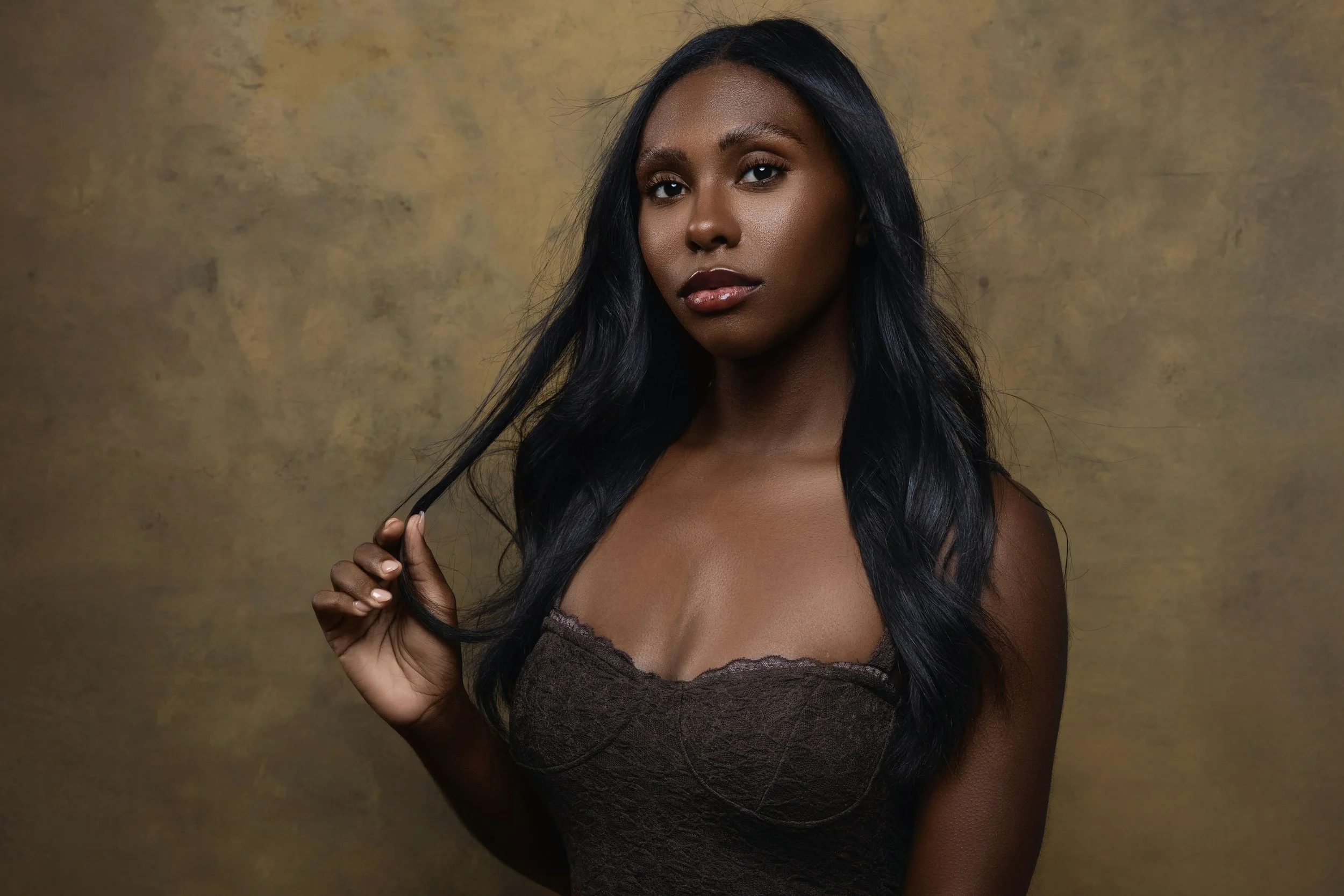 A woman with long black hair and dark skin, wearing a brown lace top, holding a strand of her hair, standing in front of a textured yellow-brown background.