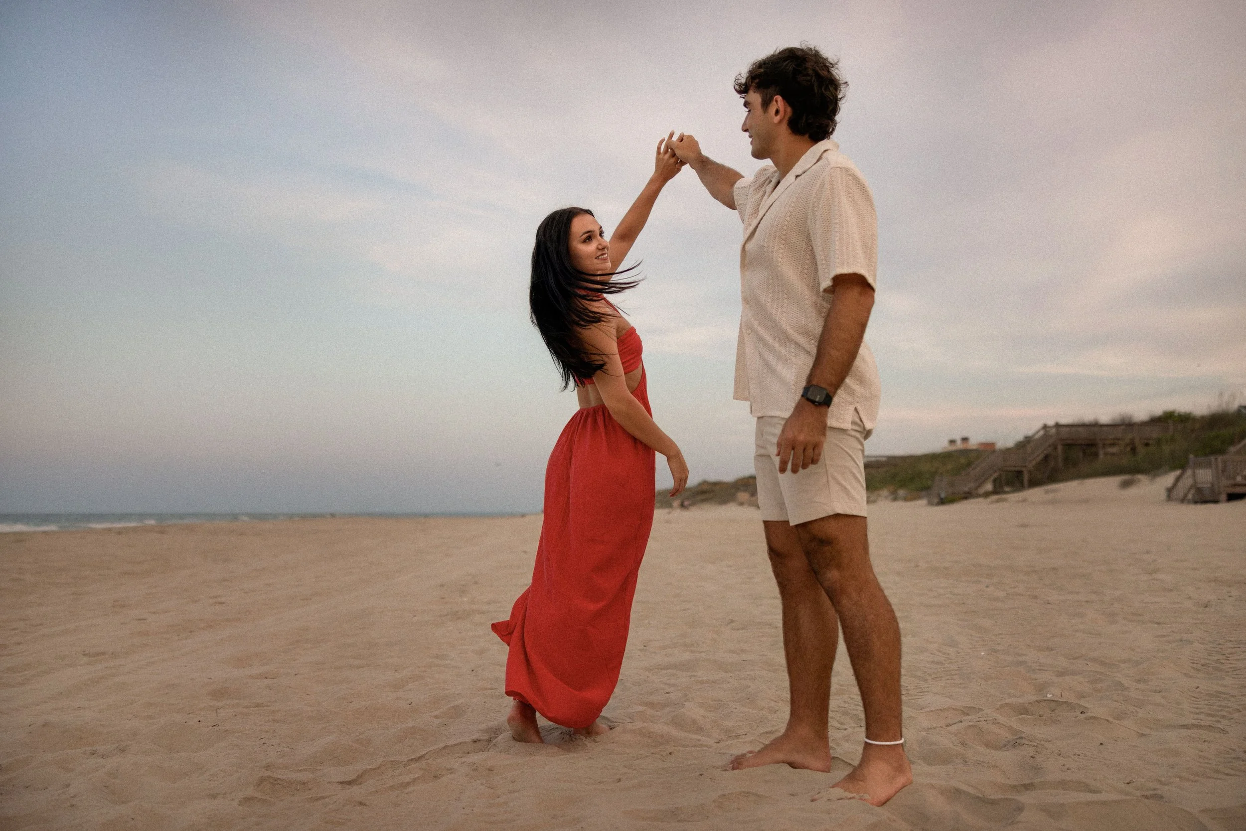 A woman in a red dress and a man in beige shorts and a light shirt dancing and holding hands on the beach during sunset.
