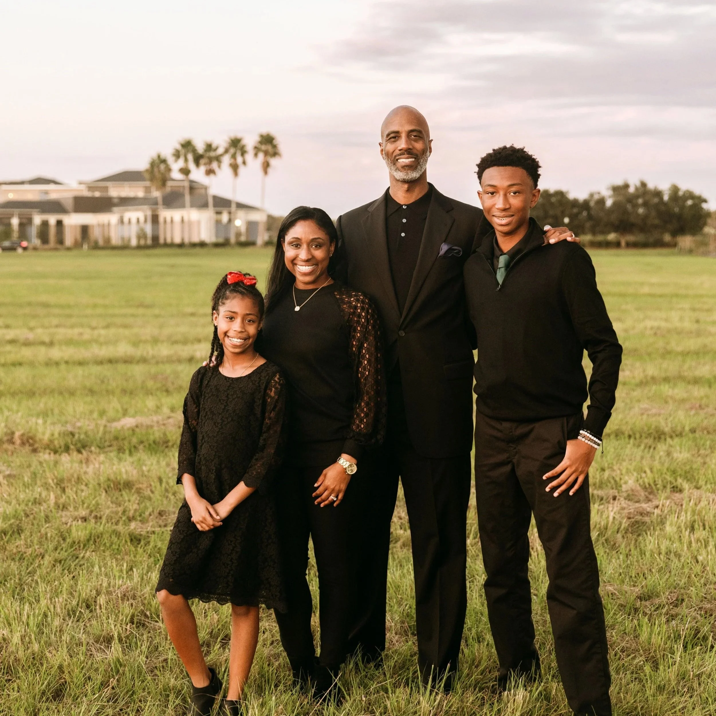 A family of five standing outdoors on a grassy field with trees and a house in the background. They are dressed in formal and semi-formal attire, smiling at the camera.