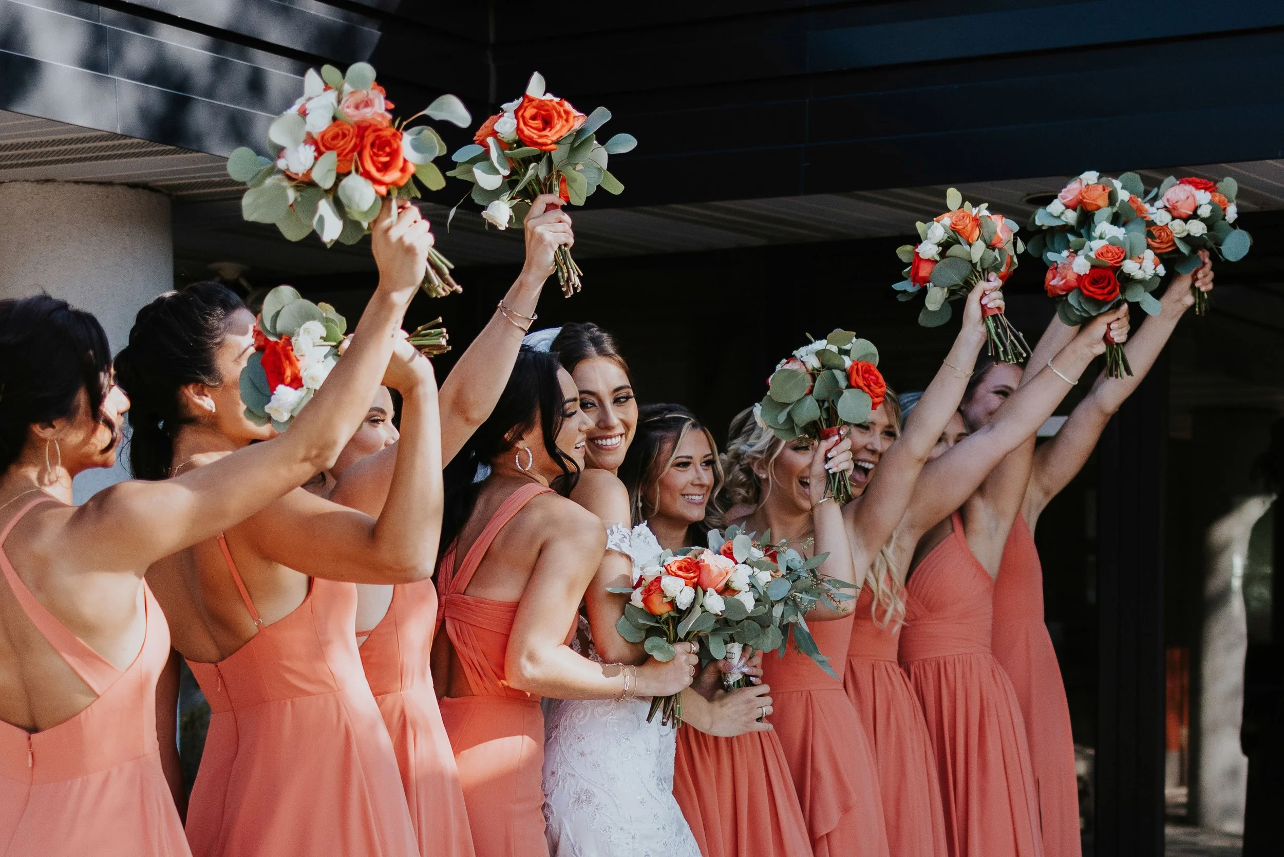 A group of women in coral dresses holding bouquets of orange and white roses with greenery, celebrating outdoors.