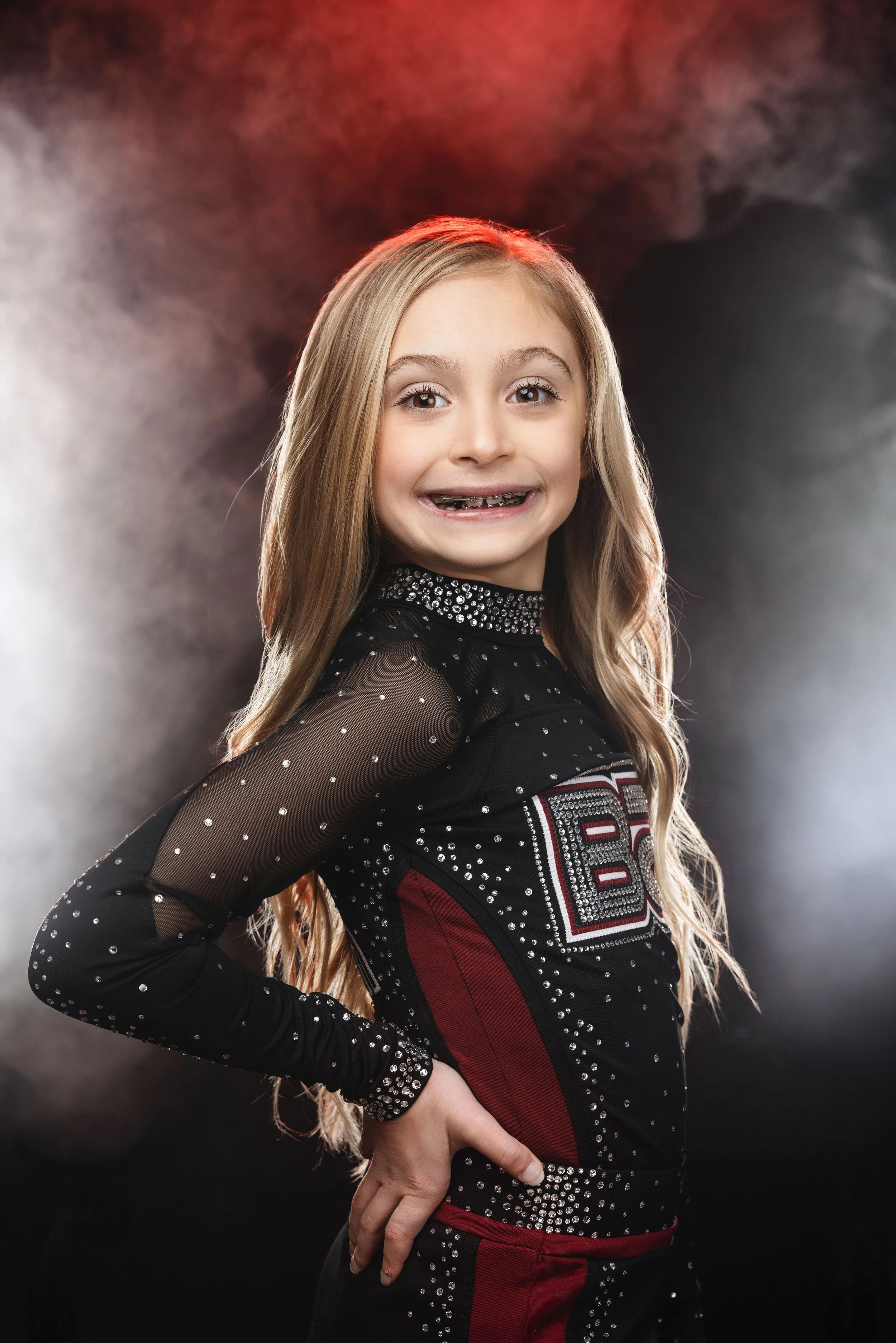 A young girl smiling with braces, wearing a black and red cheerleading uniform with rhinestones, standing against a smoky dark background.