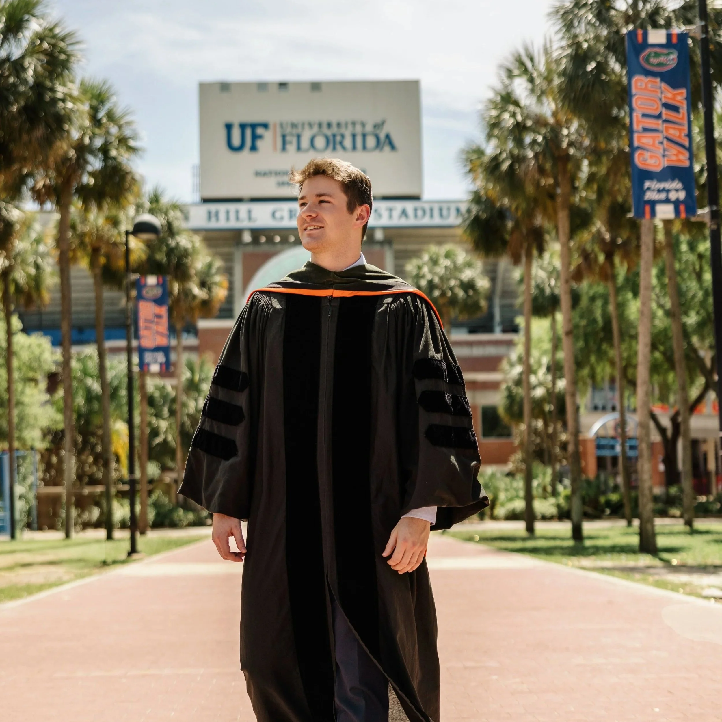 A young man in a graduation gown standing outdoors on a university campus with palm trees, a sidewalk, and a sign in the background that reads 'University of Florida.'