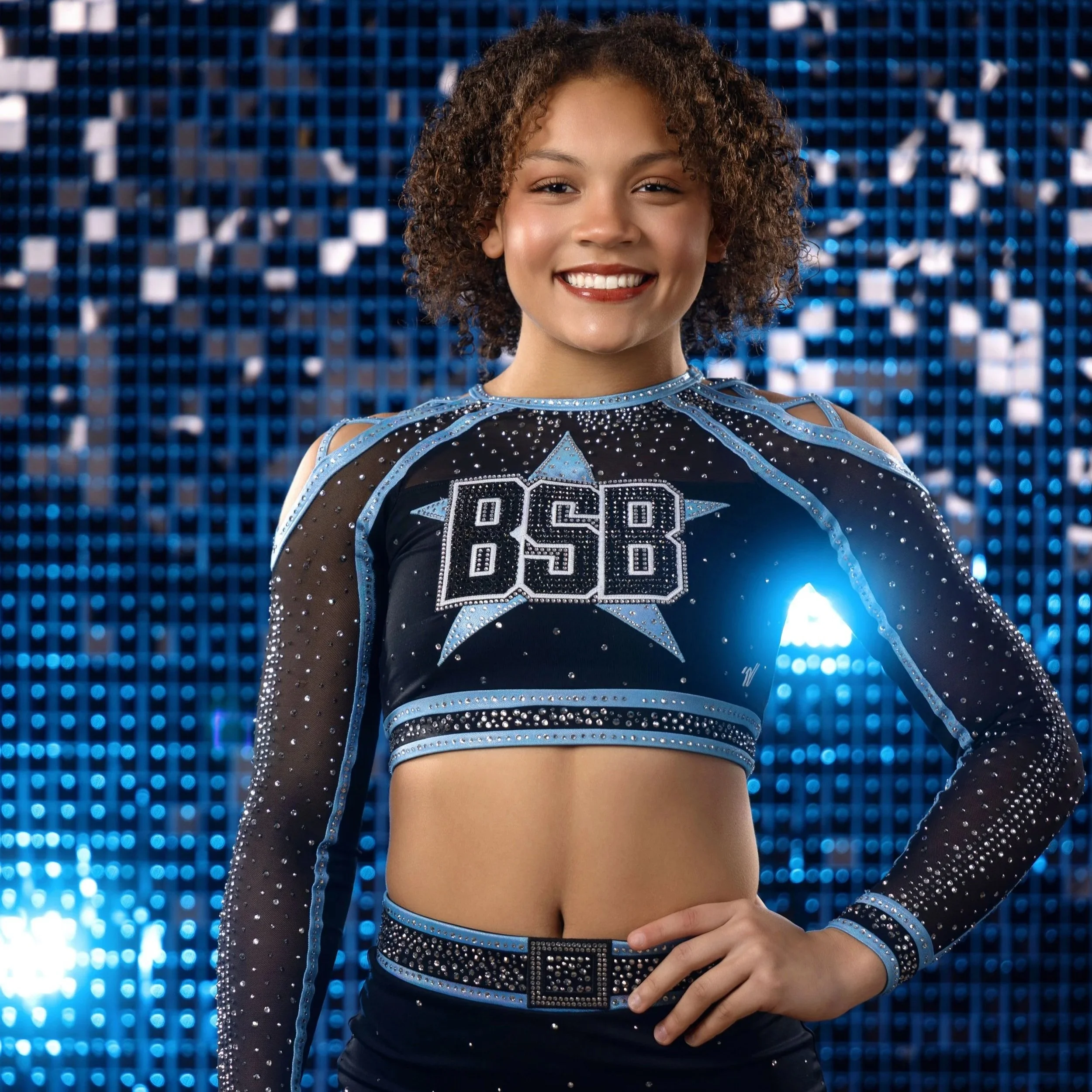 A young female cheerleader with curly hair smiling, wearing a black and blue cheerleading uniform with the number 88 on it, standing in front of a shiny blue mosaic background.