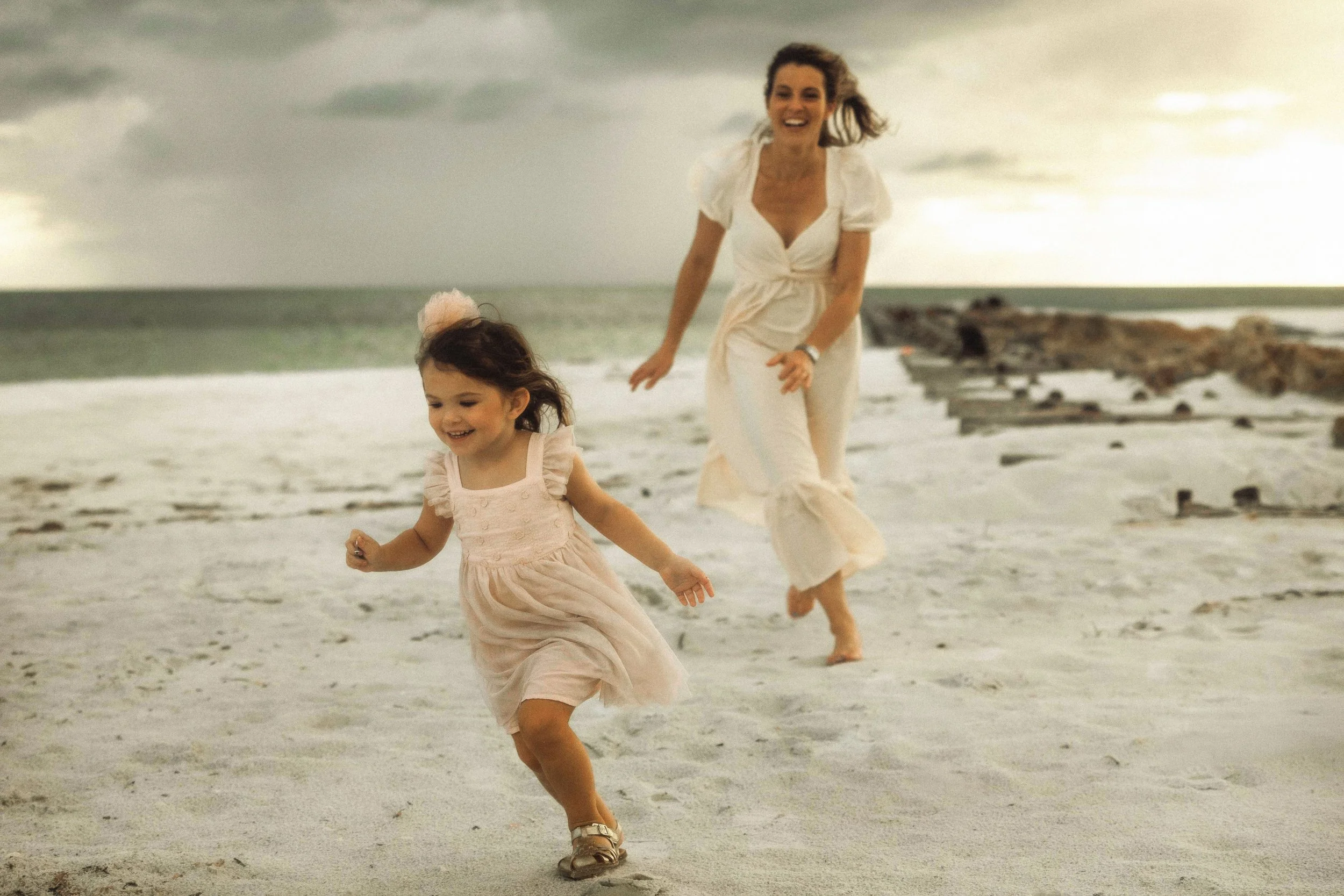 A woman and a young girl running on a sandy beach, smiling, with a cloudy sky and a rocky shoreline in the background.