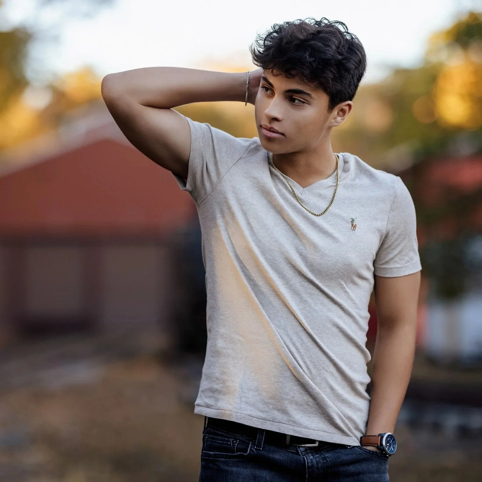 A young man with dark curly hair wearing a light gray t-shirt, gold chain, and watches, standing outdoors with blurred autumn trees in the background, touching the back of his head with one hand.