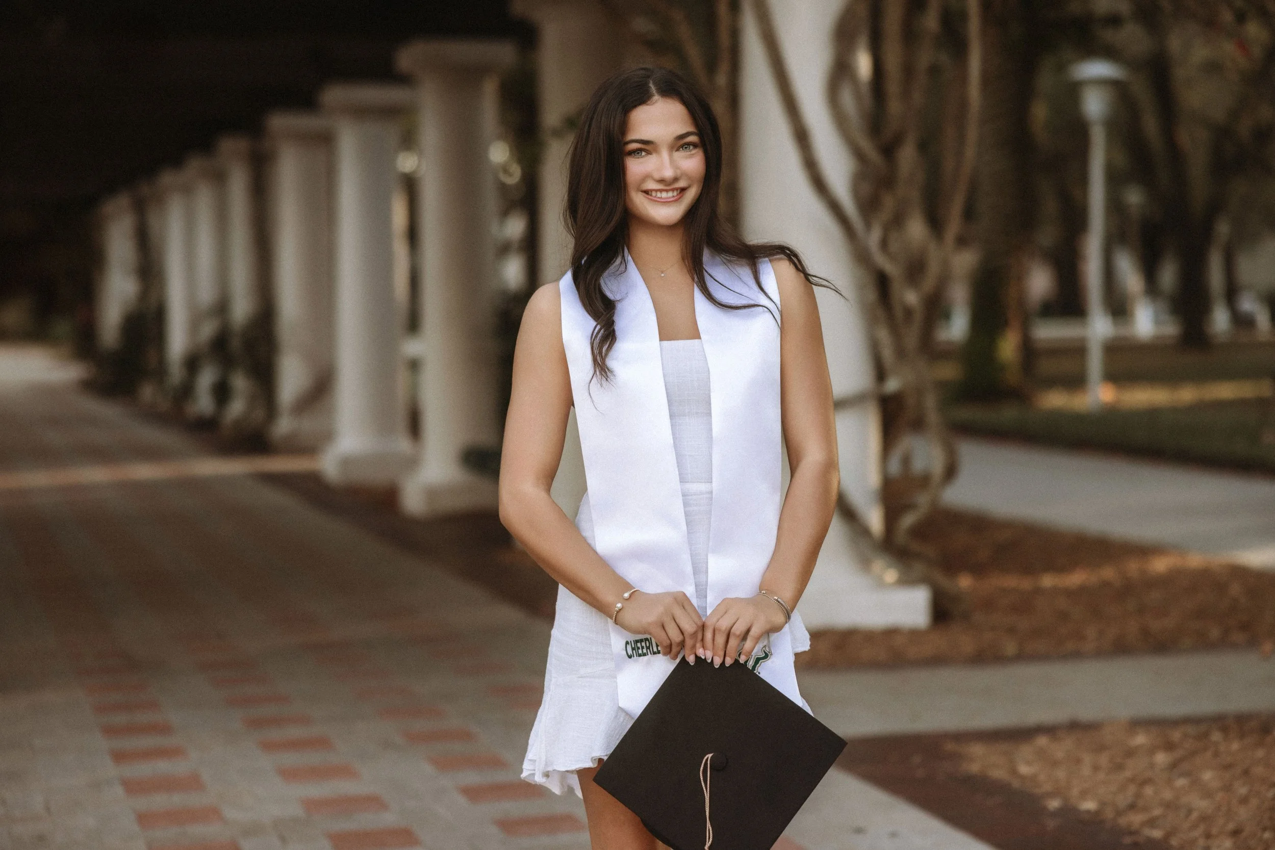 A young woman in a white dress standing outdoors near a sidewalk, smiling and holding a black graduation cap.