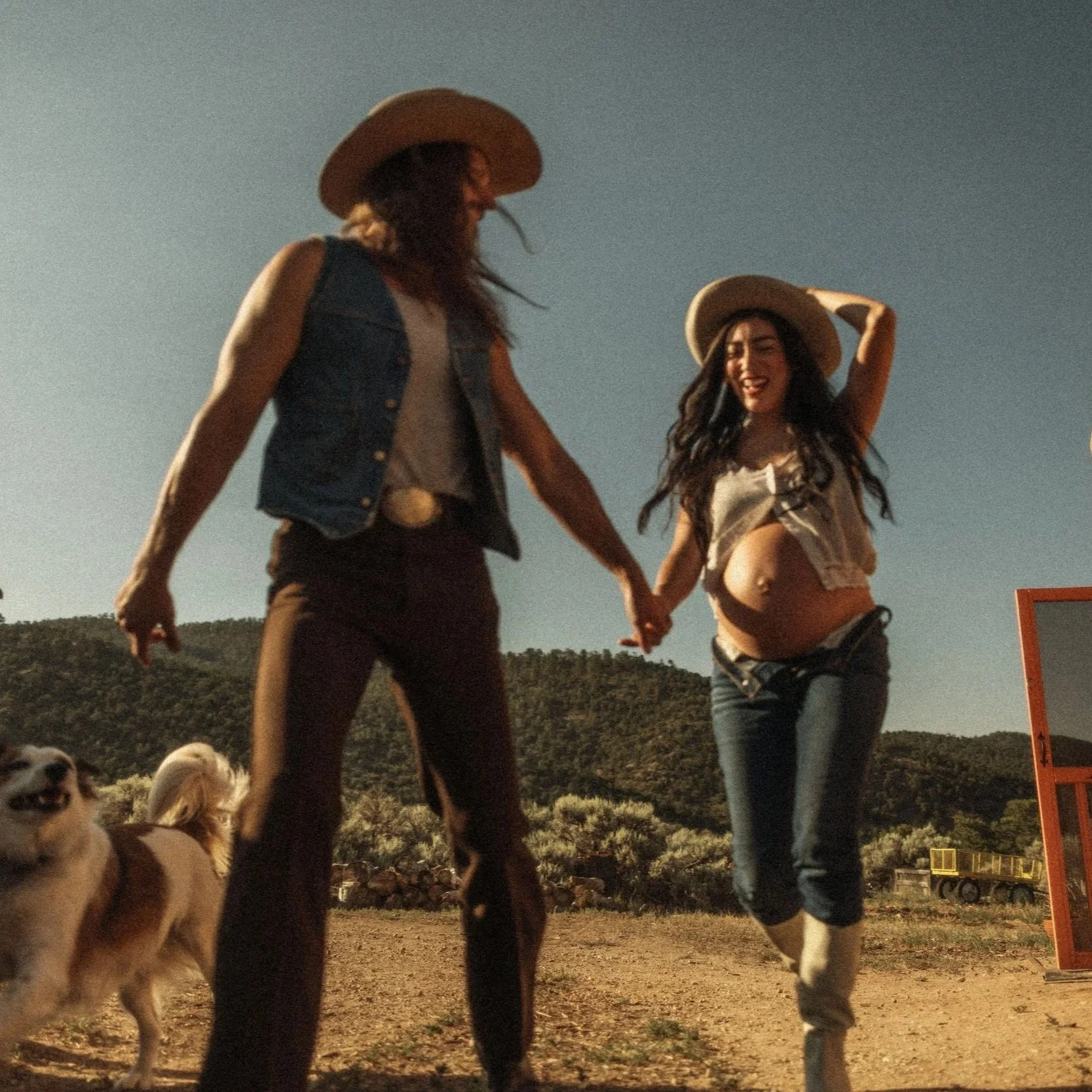 Two women holding hands and walking together outdoors during dusk, wearing cowboy hats and casual clothing, with dogs nearby and a scenic landscape in the background.