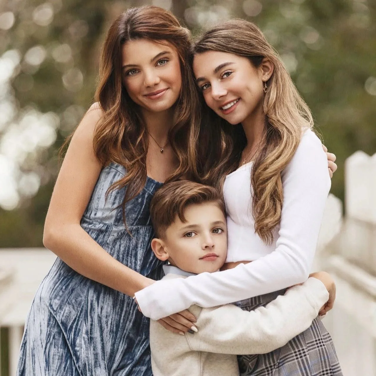 Three females and one young boy standing closely together outdoors, smiling softly, with trees in the background.