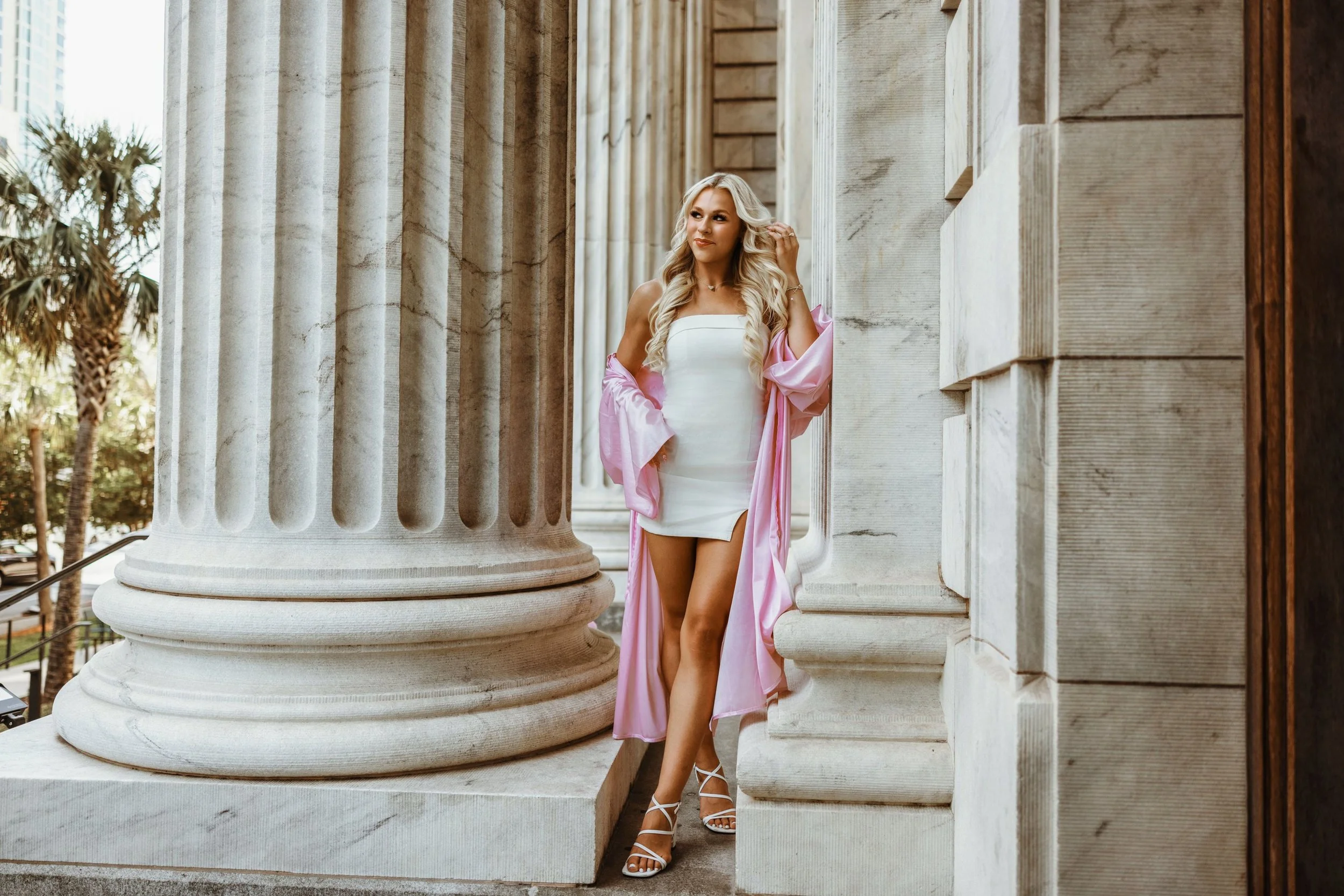 A woman with long blonde hair in a white dress and pink satin robe stands between large stone columns of a neoclassical building, looking to her left with a slight smile.
