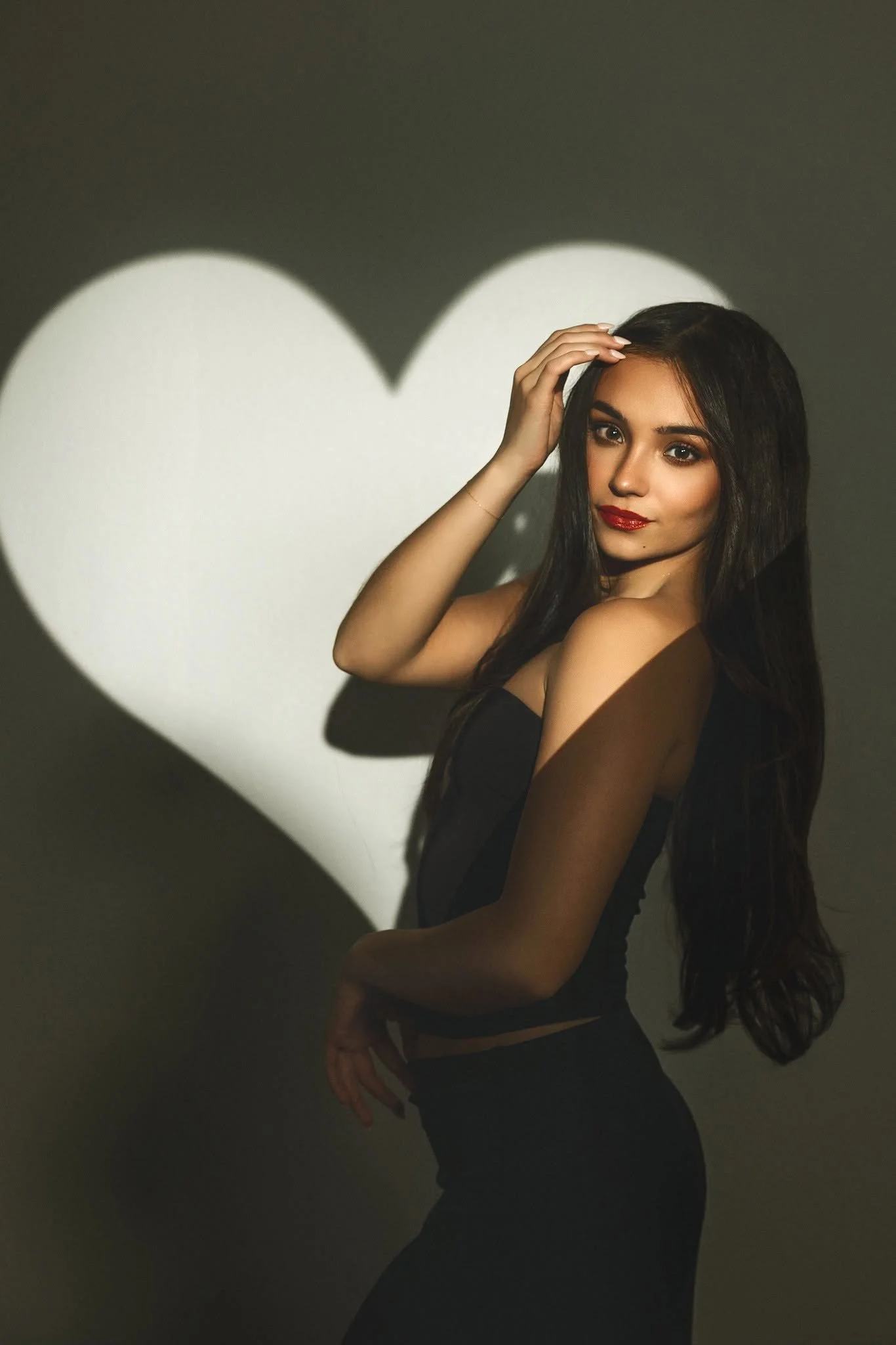 A woman with long dark hair and makeup, wearing a black strapless dress, posing in front of a dark wall with a large white heart-shaped shadow projected behind her.
